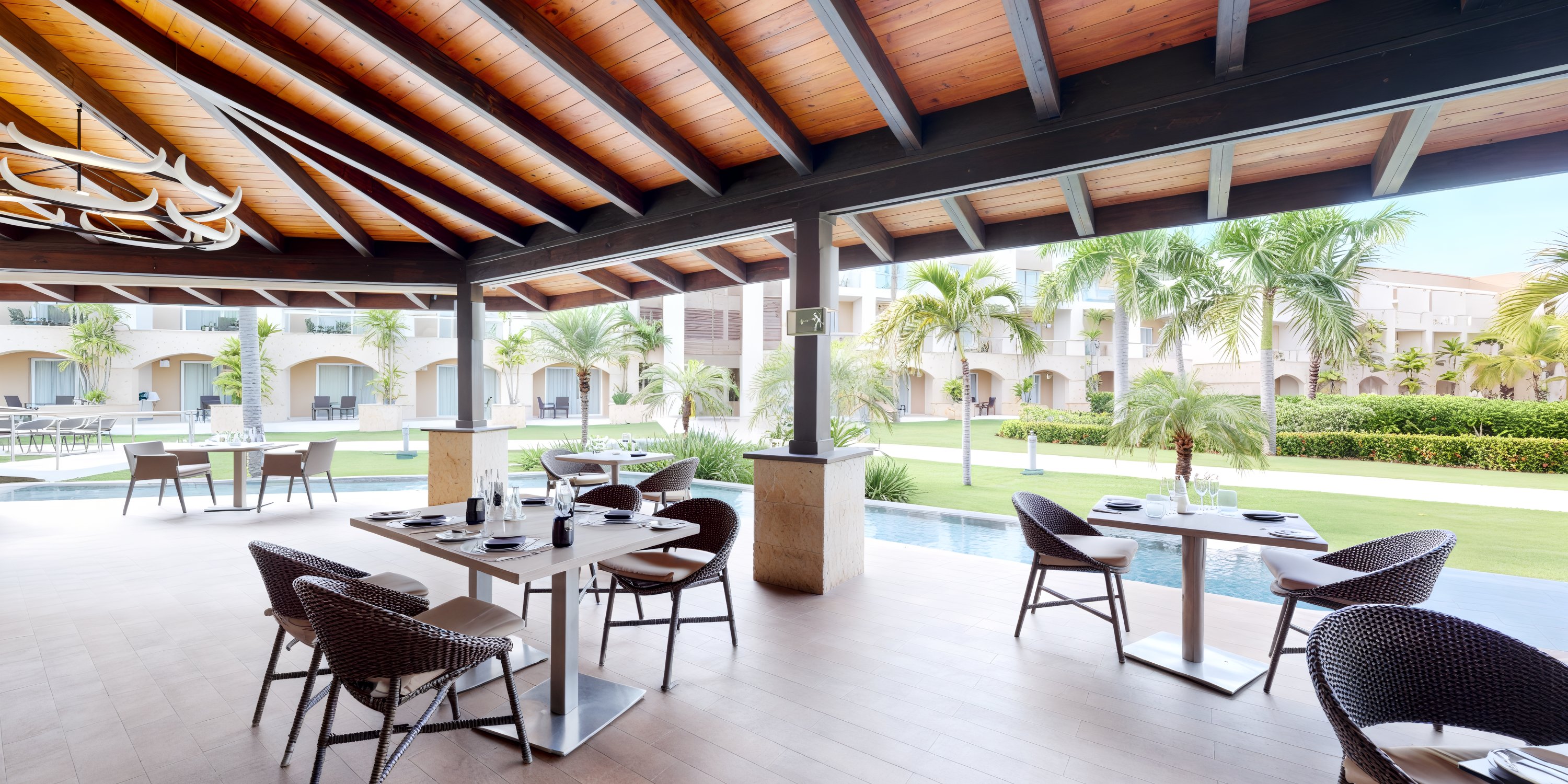 An outdoor dining area featuring tables and chairs beneath a wooden roof offers the perfect setting for a destination wedding, with scenic views of a lush garden adorned with palm trees.