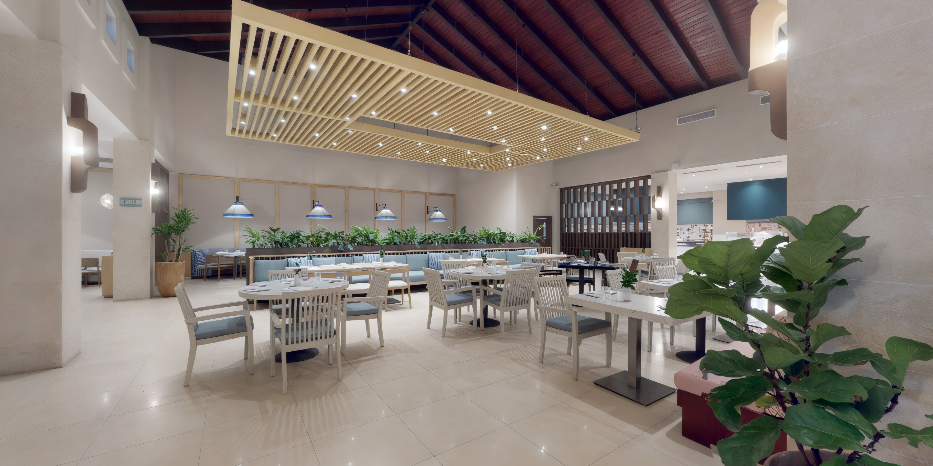 A contemporary restaurant interior, perfect for a destination wedding reception, features white tables and chairs elegantly arranged among lush green plants beneath a warm wooden ceiling.