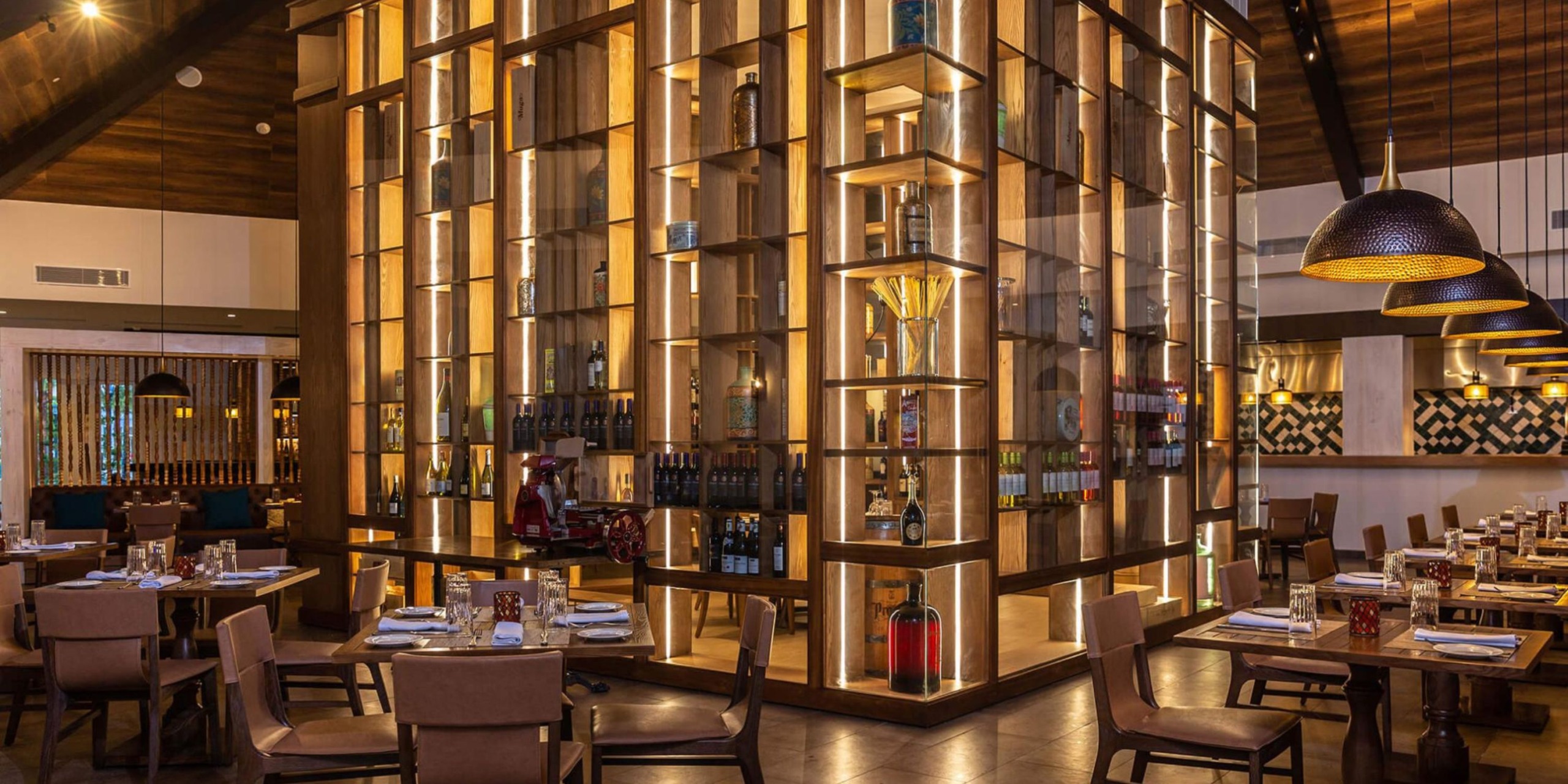 The Margaritaville Island Reserve Riviera Maya features a chic restaurant interior with wooden decor, a centralized bottle display, tables elegantly set with dinnerware, and inviting warm lighting.