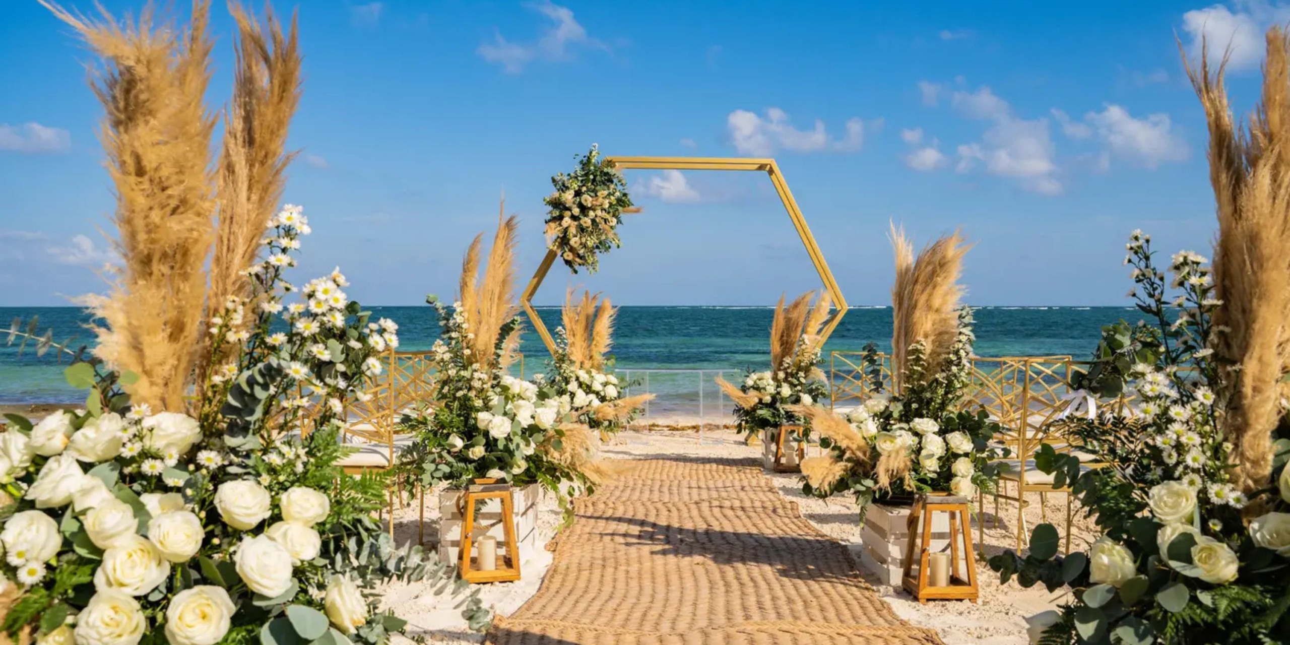 The Margaritaville Island Reserve Riviera Maya offers a beachfront wedding setup featuring a hexagonal arch with greenery and white flowers. Pampas grass and roses line the sandy aisle, leading to the ocean under a serene blue sky.