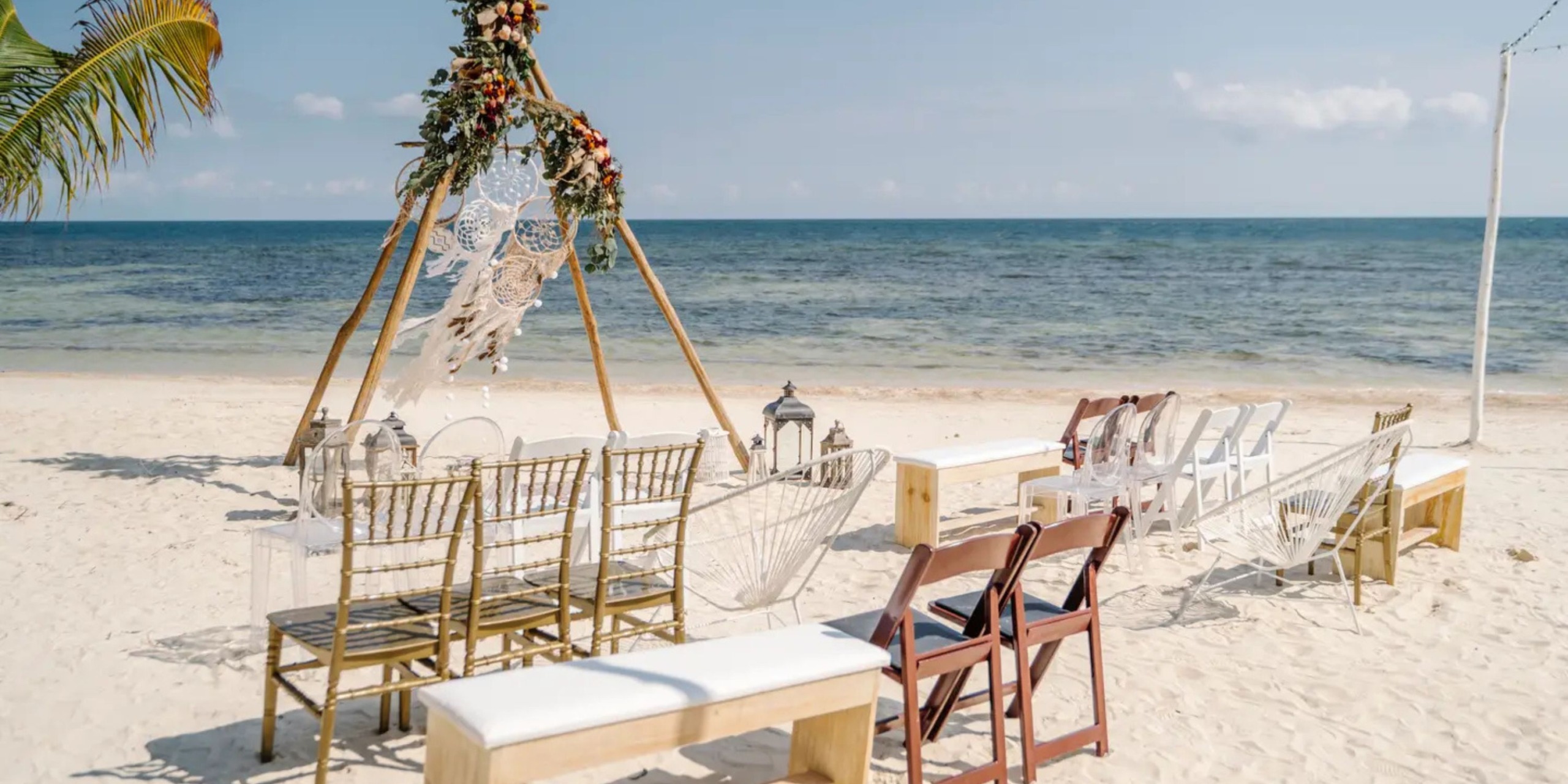 A beach wedding setup at Margaritaville Island Reserve Riviera Maya features chairs in rows facing an altar adorned with macramé and flowers, set against the backdrop of ocean waves and a blue sky.