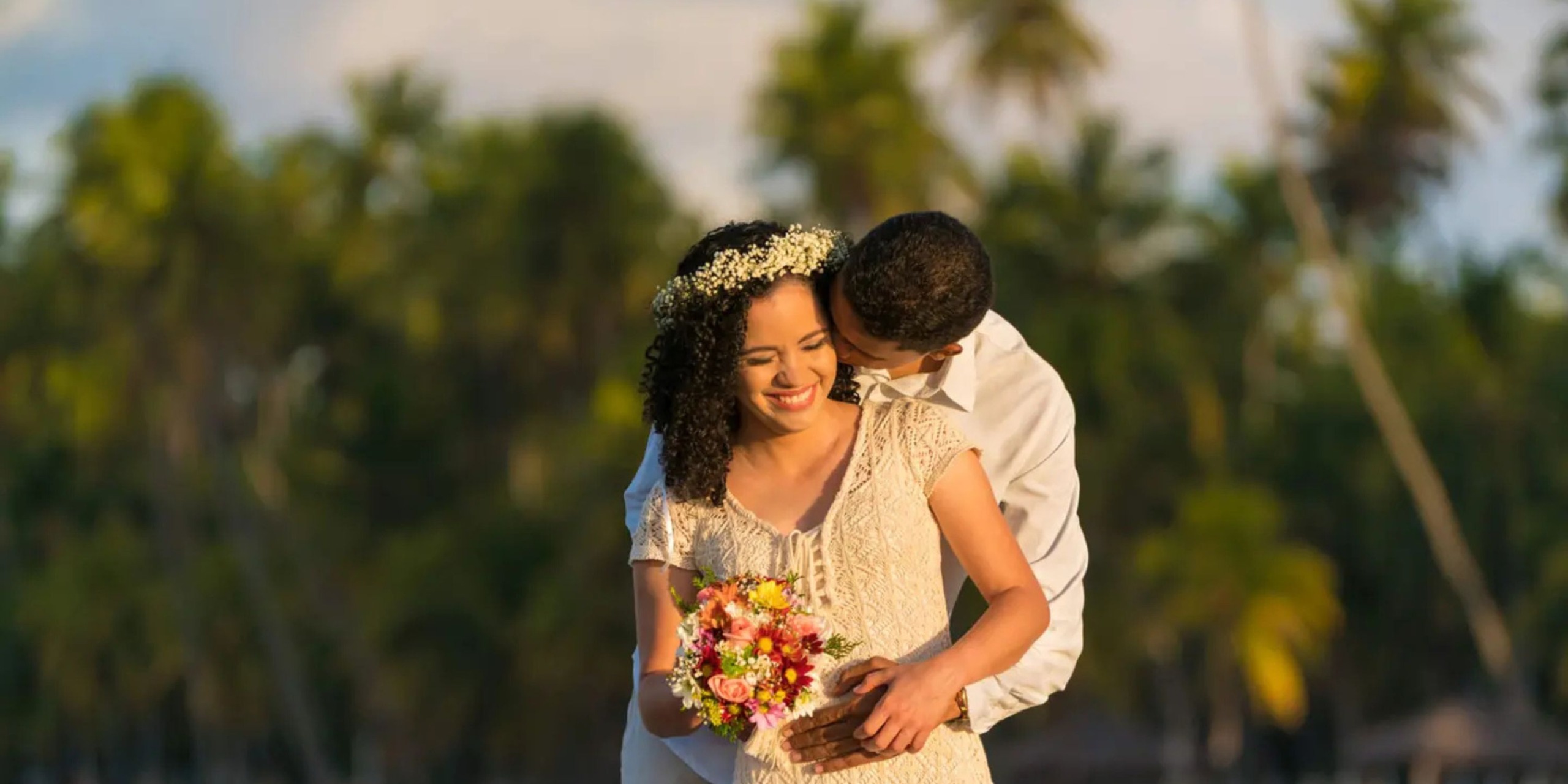 A couple embraces happily in the tropical paradise of Margaritaville Island Reserve Riviera Maya, with the woman holding a vibrant bouquet. Palm trees stand majestically in the background.