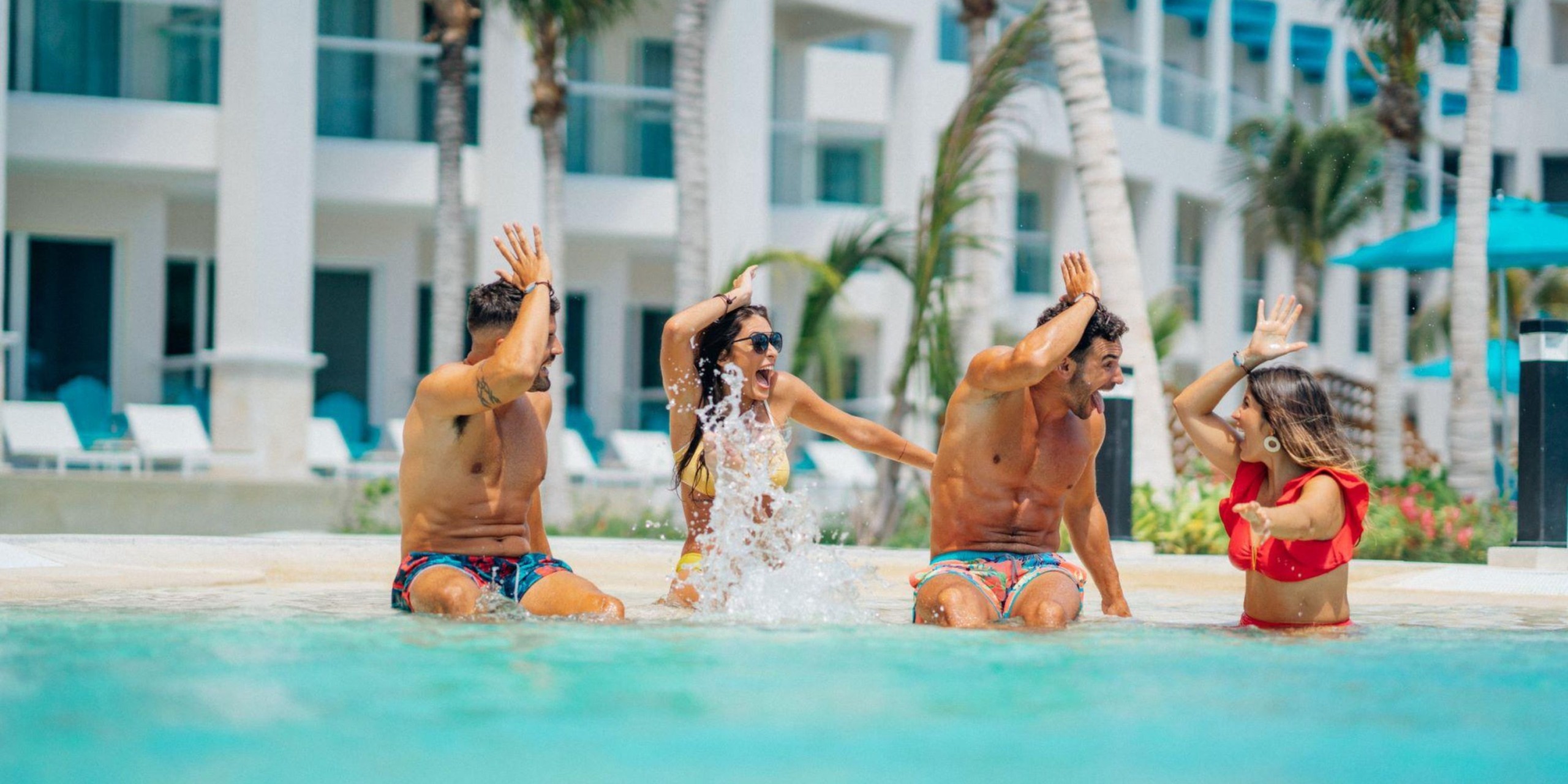 Four people in swimwear splash water playfully in the pool at Margaritaville Island Reserve Riviera Maya, surrounded by a resort scene with palm trees and white buildings.
