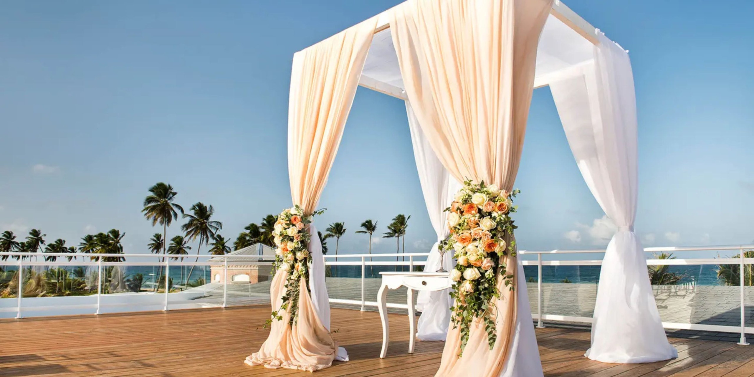 The Margaritaville Island Reserve Riviera Maya features an elegant wedding arbor adorned with draped fabric and floral arrangements, set on a sunny beachside terrace with palm trees and an ocean view.