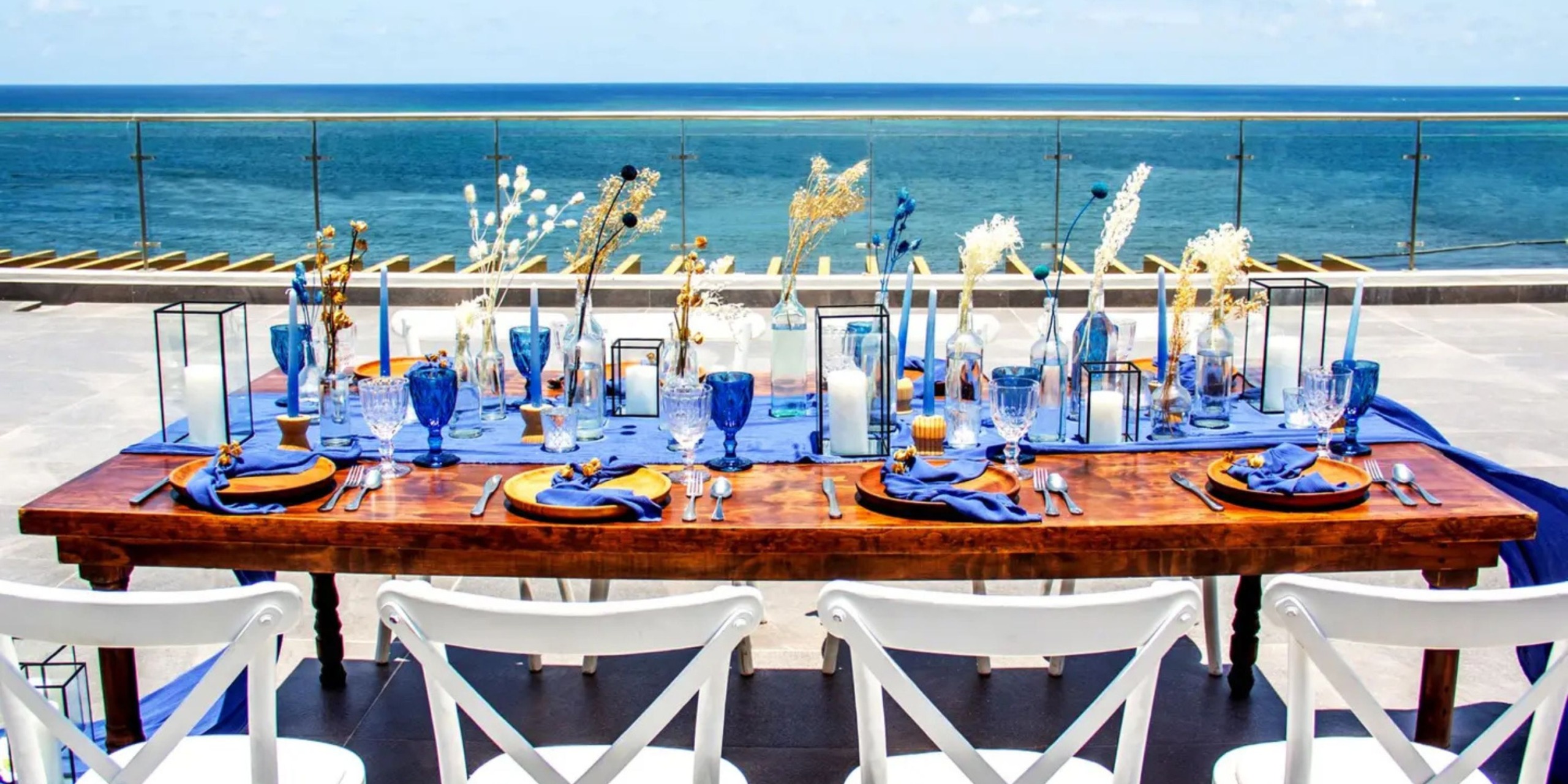 The Margaritaville Island Reserve Riviera Maya features a wooden dining table for six adorned with blue glassware, orange napkins, and decorative vases on a terrace overlooking an ocean under a clear sky.
