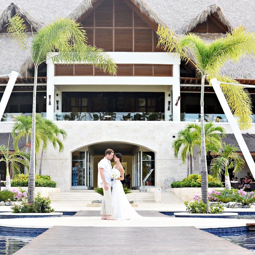 A bride and groom standing in front of a resort with thatched roofs.