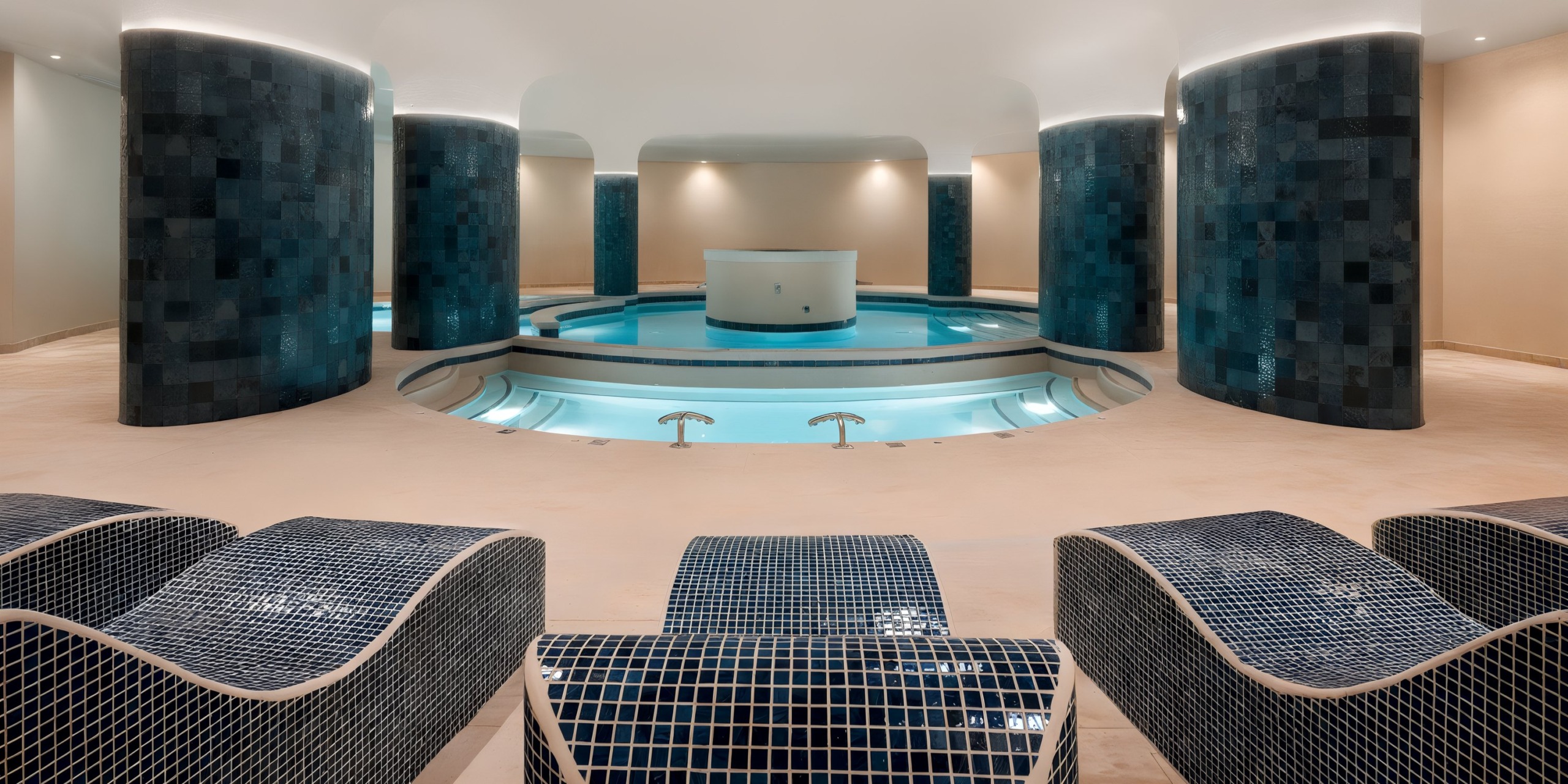 Discover the expansive indoor spa at AVA Resort Cancun, showcasing multiple blue-tiled pools surrounded by elegant tall curved columns and tiled lounge chairs in the foreground.