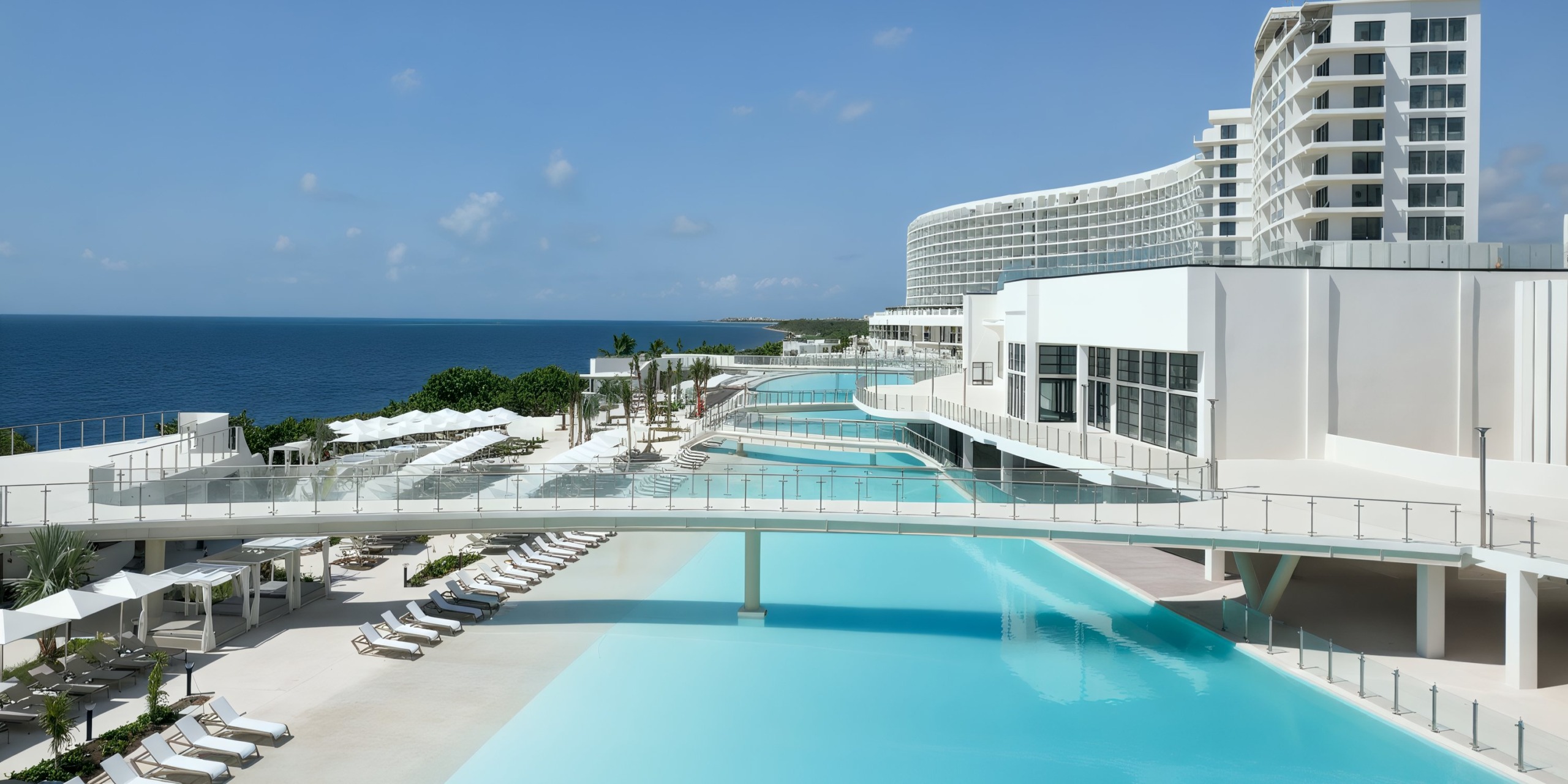 AVA Resort Cancun features a large pool, sun loungers, and modern white buildings overlooking the ocean under a clear blue sky.