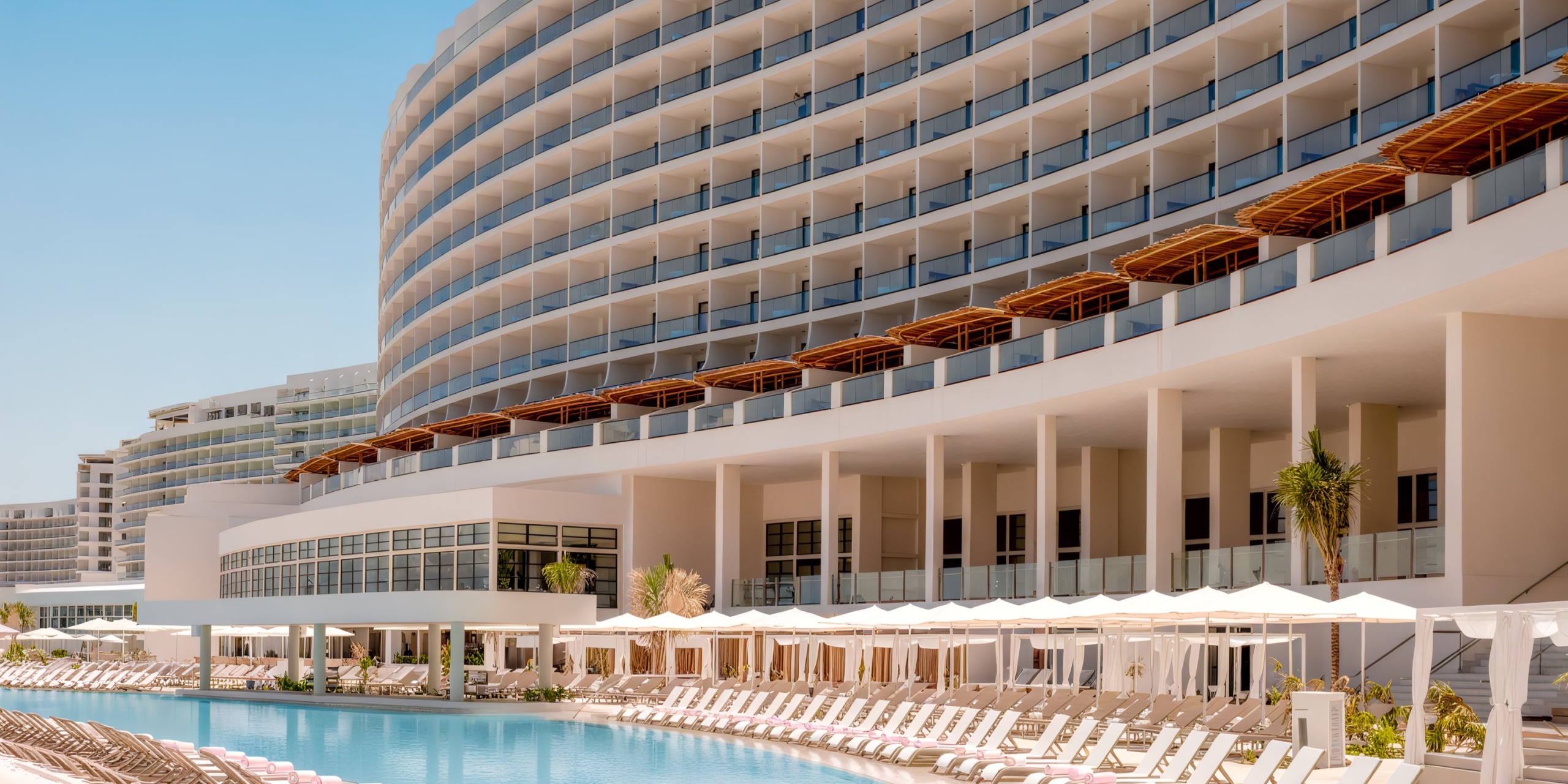 The AVA Resort Cancun features a contemporary curved design with balconies that offer views of a spacious pool, lined with sun loungers and umbrellas beneath a clear blue sky.