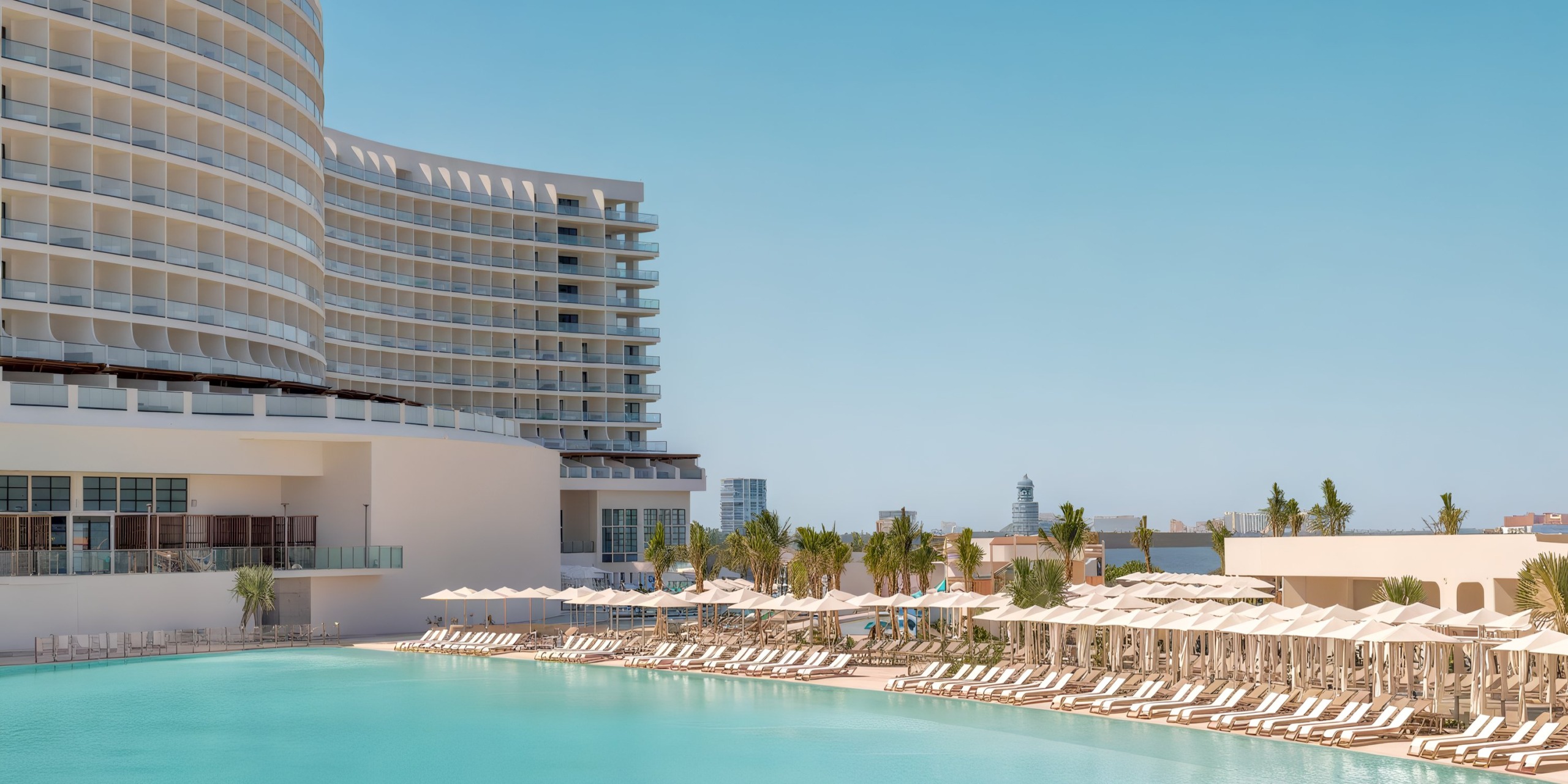 AVA Resort Cancun is a spacious, contemporary resort with distinctive curved architecture that offers stunning views of an expansive outdoor pool, adorned with rows of white loungers and umbrellas set against a clear blue sky.