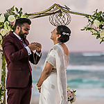 A bride and groom exchanging rings under a floral arch on a beach, with the ocean in the background.