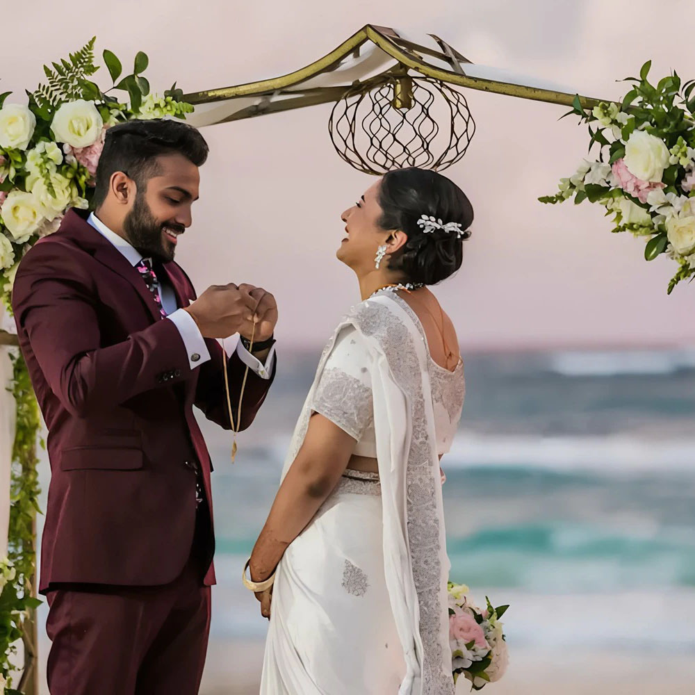 A bride and groom exchanging rings under a floral arch on a beach, with the ocean in the background.