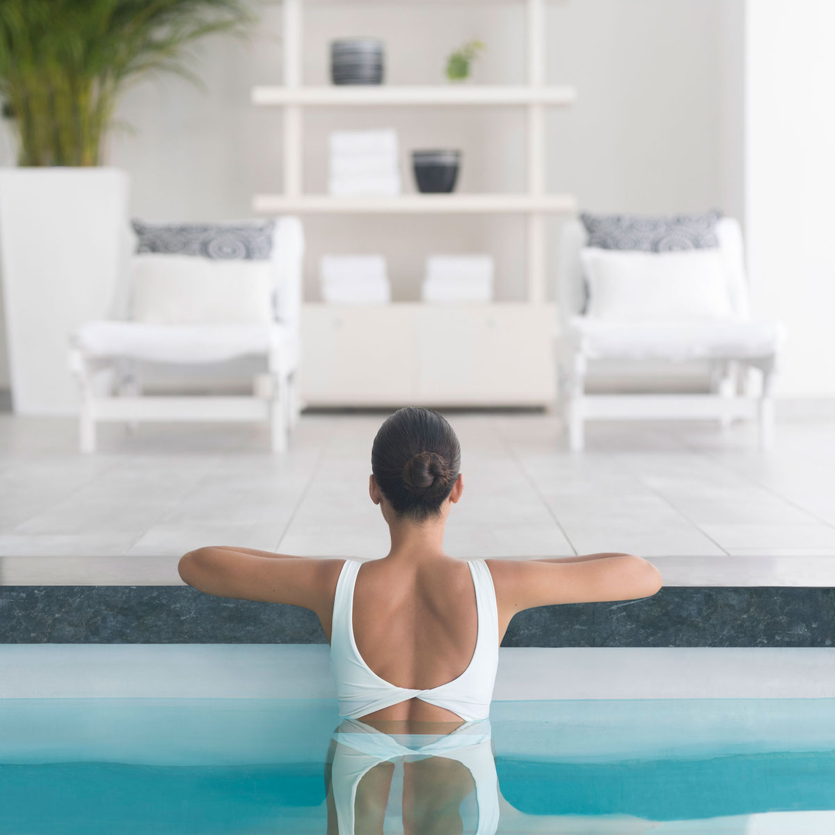 A woman in a white swimsuit relaxes by an edge of a pool, facing away, looking towards a bright, stylishly decorated room at a destination wedding.