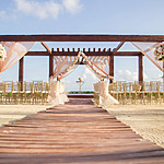 A beach wedding setup with a wooden aisle leading to a flower-adorned arch with elegant wedding draping and lined with chairs under a clear blue sky.