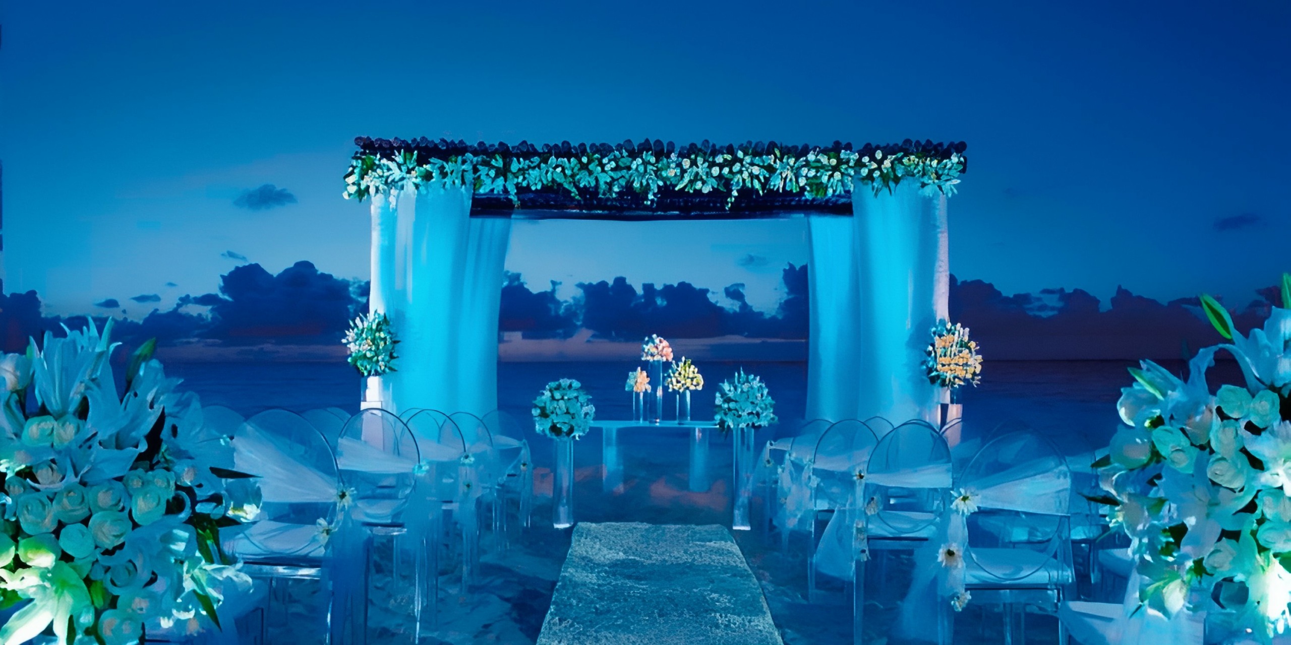 A beachside wedding setup with white flowers, transparent chairs, and an aisle leading to a floral arch at Dreams Estrella del Mar Mazatlan, all under a twilight sky.
