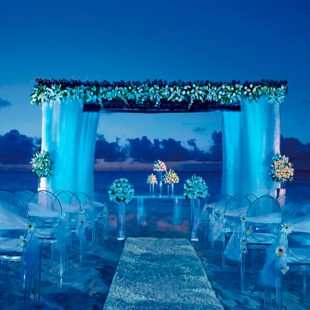 A beach wedding setup at dusk with an altar decorated with flowers and blue drapery, clear chairs with white sashes, and a flower-lined aisle leading to the altar.