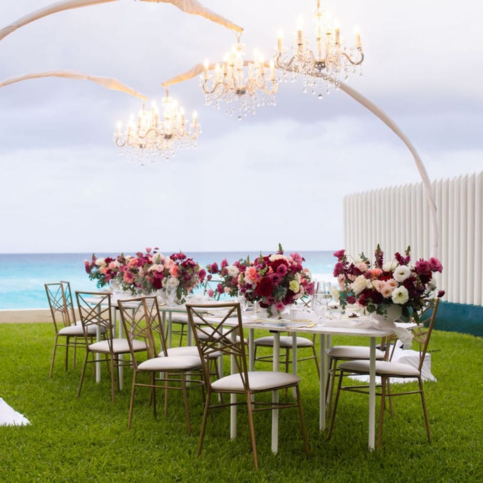 Destination wedding outdoor dining table adorned with floral centerpieces, chandeliers, and ocean view.