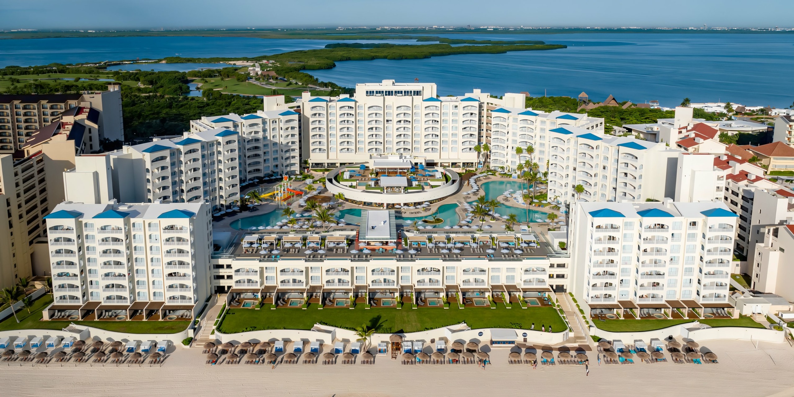 Aerial view of Hilton Cancun Mar Caribe, a large beachfront resort with multiple buildings, swimming pools, and sun loungers, adjacent to a turquoise sea and surrounded by lush greenery.