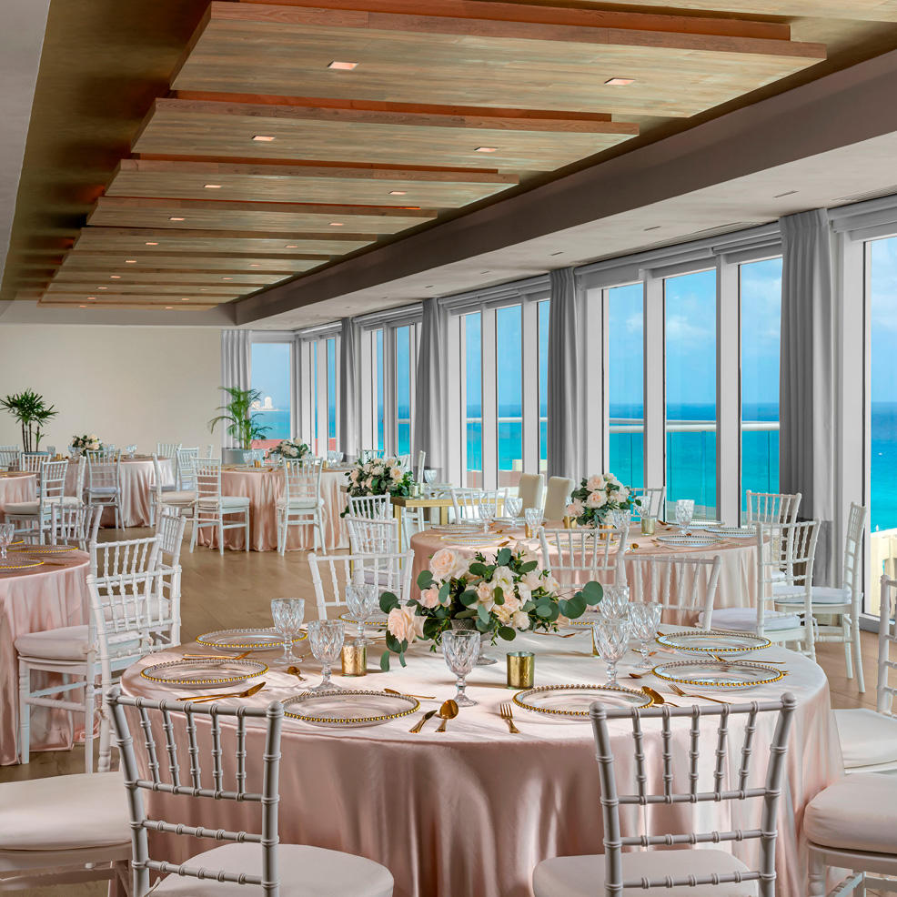 Elegant dining room setup for a wedding with round tables, floral centerpieces, and ocean view through large windows.