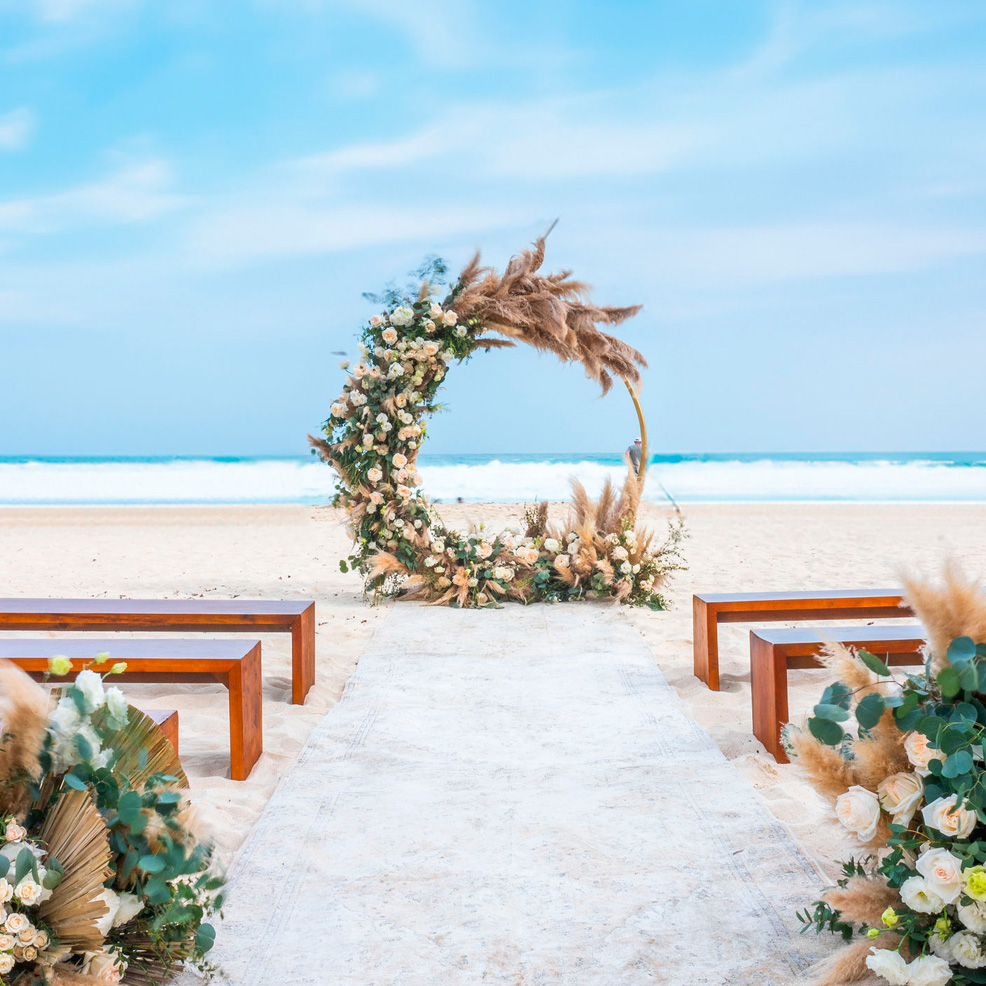 A beach wedding setup with a floral arch, a white aisle, and wooden benches, overlooking the ocean.