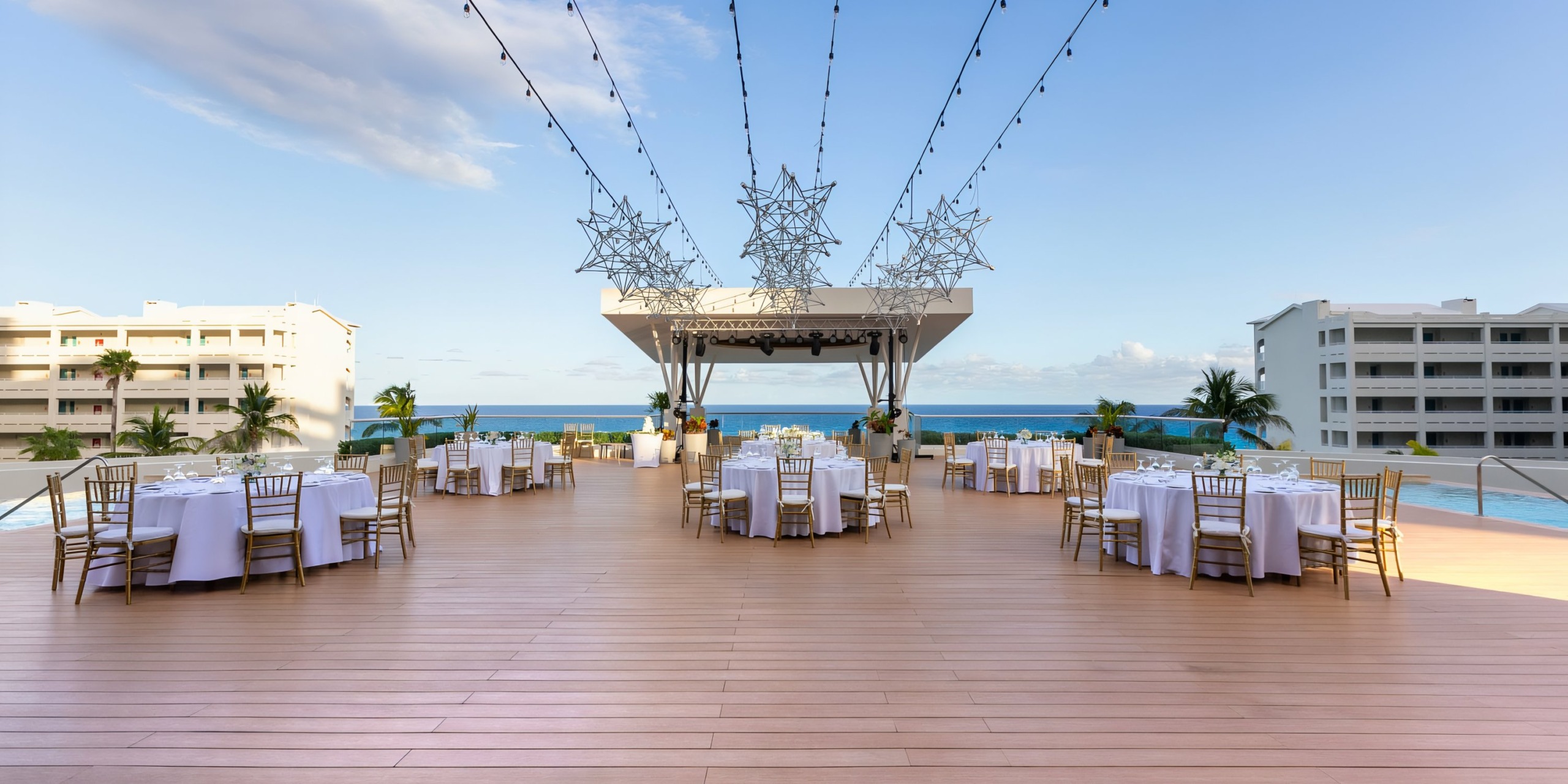Hilton Cancun Mar Caribe outdoor wedding reception area with white tables set up on a wooden deck, decorative lights overhead, and a view of the ocean and buildings in the background.