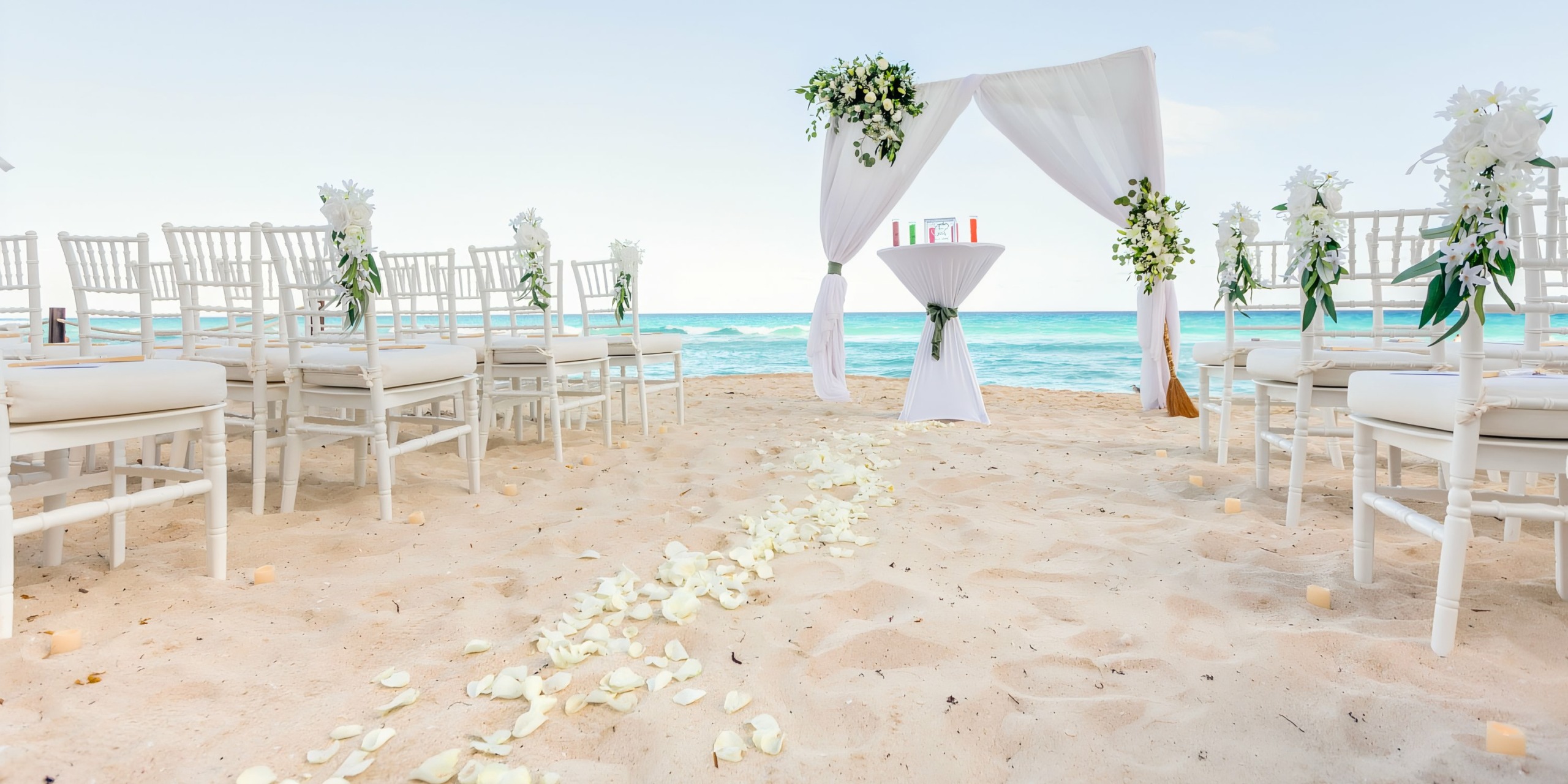 A beach wedding setup with white chairs, a floral decorated altar, and a petal-strewn aisle leading to the ocean at Hilton Cancun Mar Caribe.