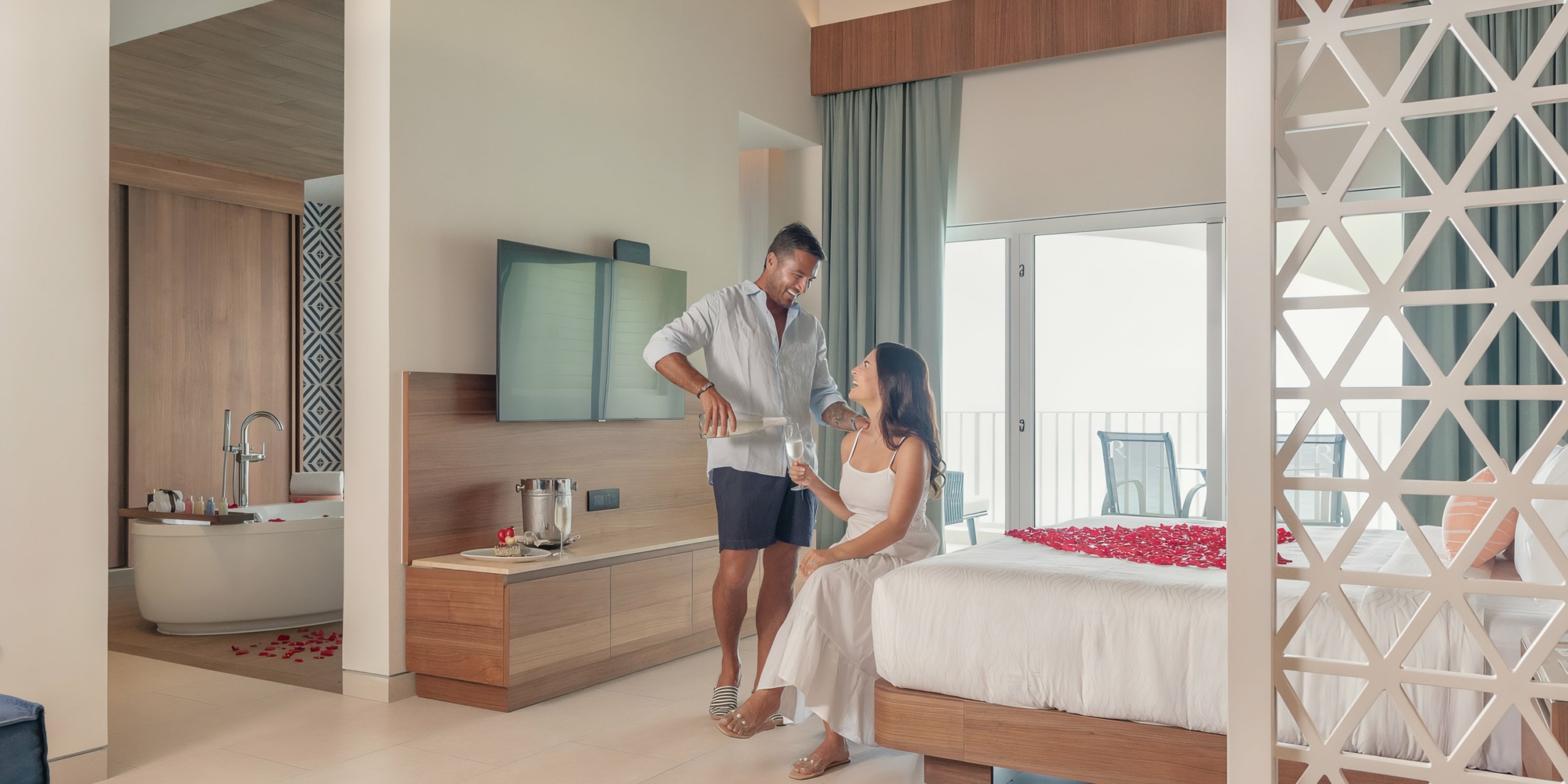 A man serving coffee to a woman sitting on the edge of a bed in the Hilton Cancun Mar Caribe with open balcony doors.