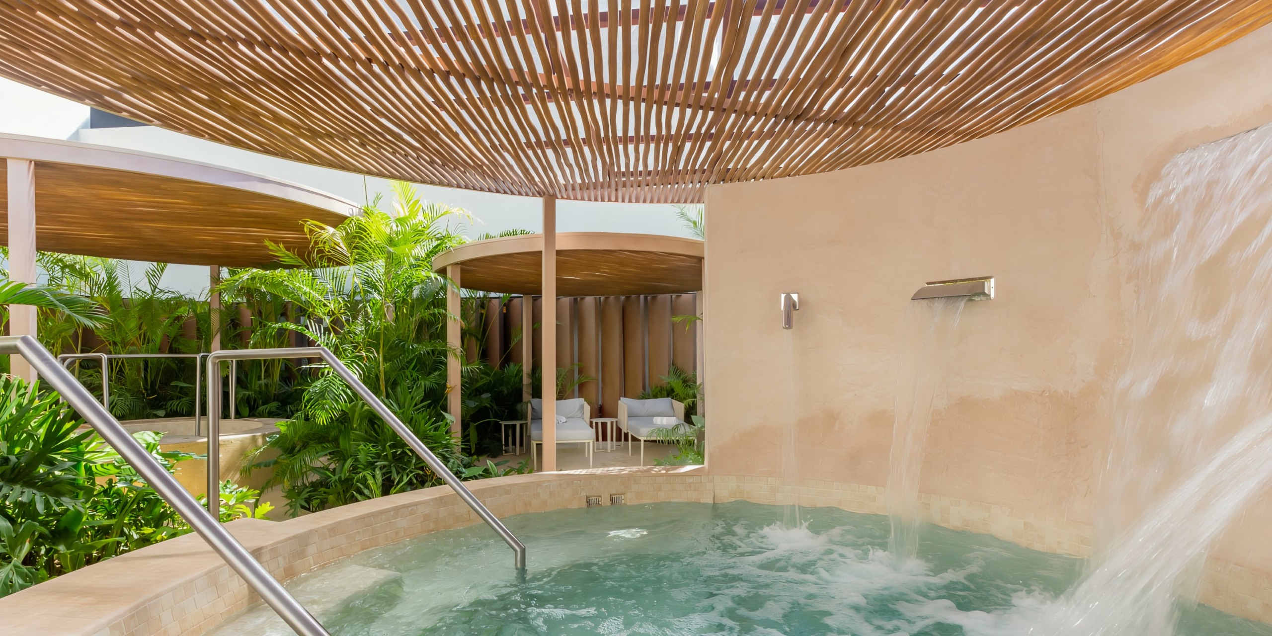 Outdoor spa area with a whirlpool and cascading water, surrounded by privacy walls and a wooden lattice roof, with lush greenery visible in the background at Hilton Cancun Mar Caribe.