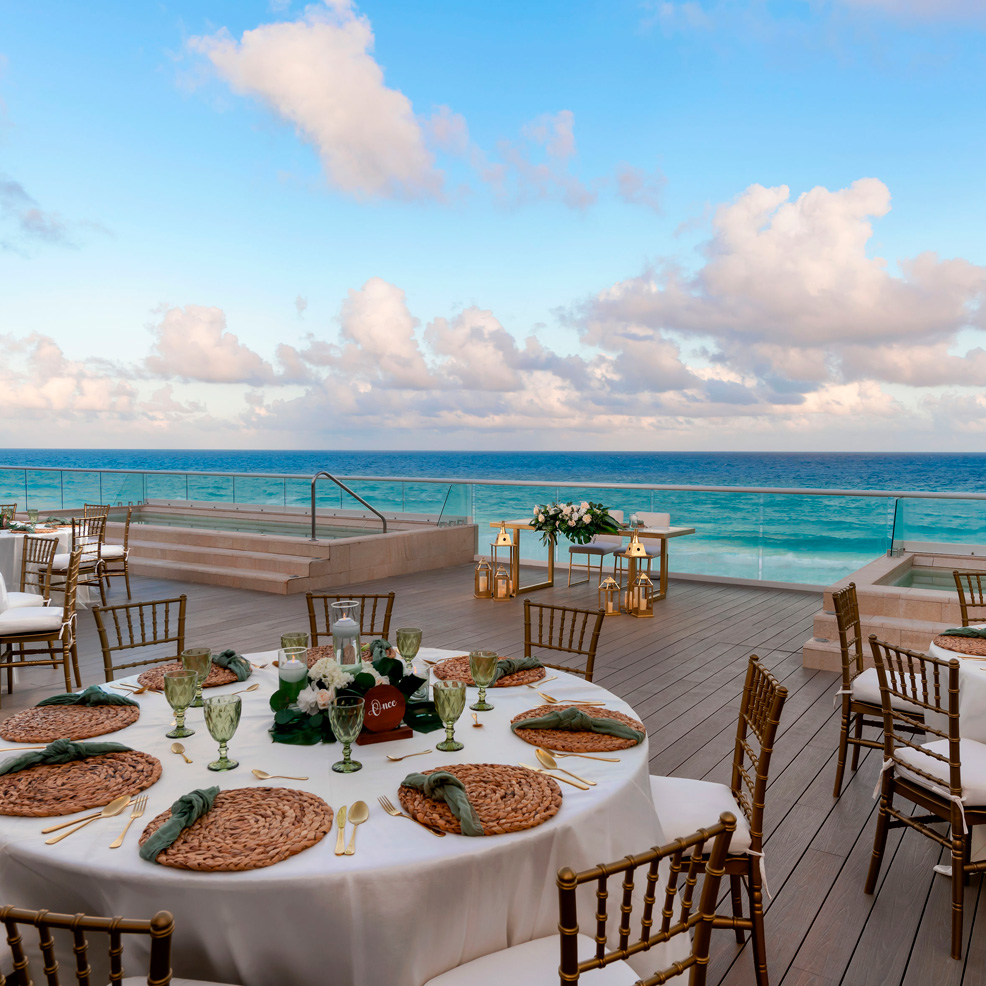 Outdoor dining setup on a wooden deck overlooking the ocean, featuring elegantly set tables with glassware and a clear blue sky above.