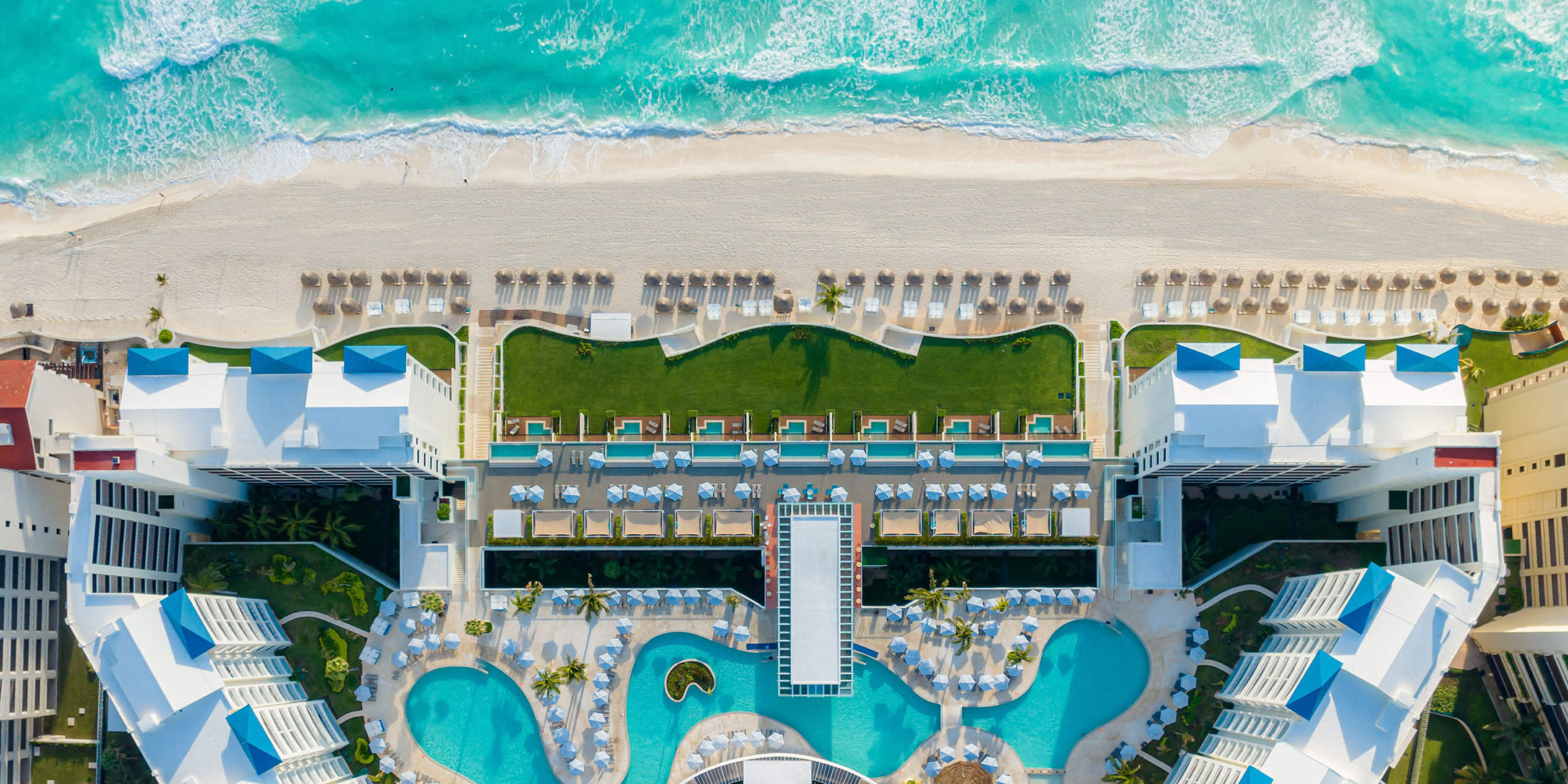 Aerial view of Hilton Cancun Mar Caribe resort with pools, umbrellas, and loungers, flanked by a turquoise ocean on one side and landscaped gardens on the other.