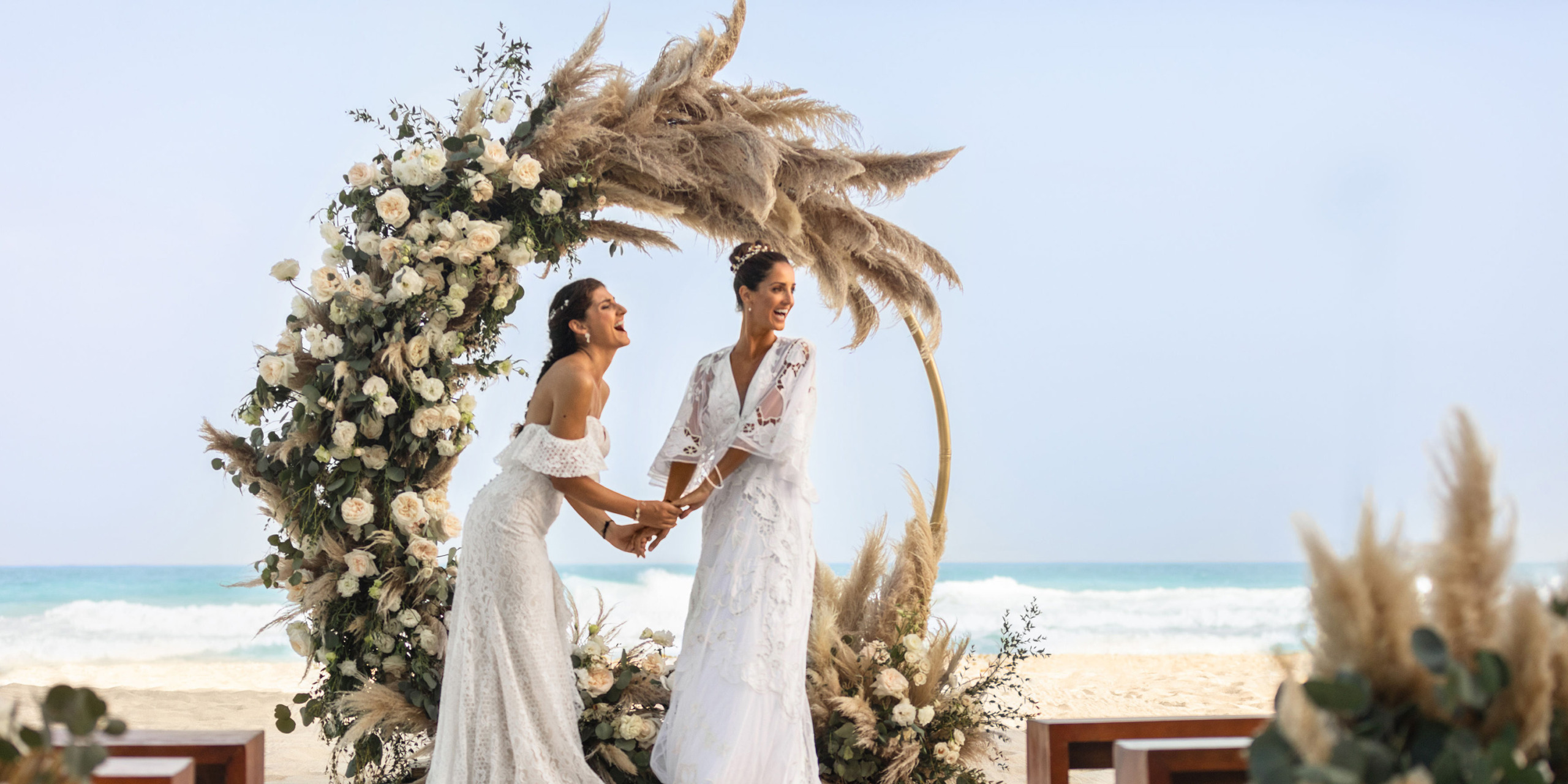 Two brides holding hands under a Hilton Cancun Mar Caribe on a beach, with the ocean in the background.