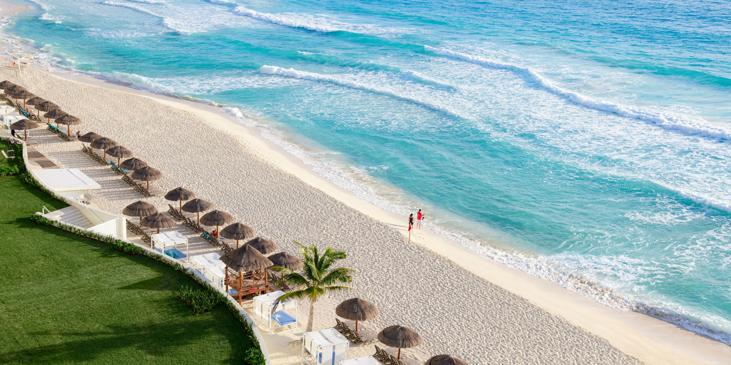 Aerial view of Hilton Cancun Mar Caribe with turquoise waves, umbrellas, and a lone person walking along the shore.
