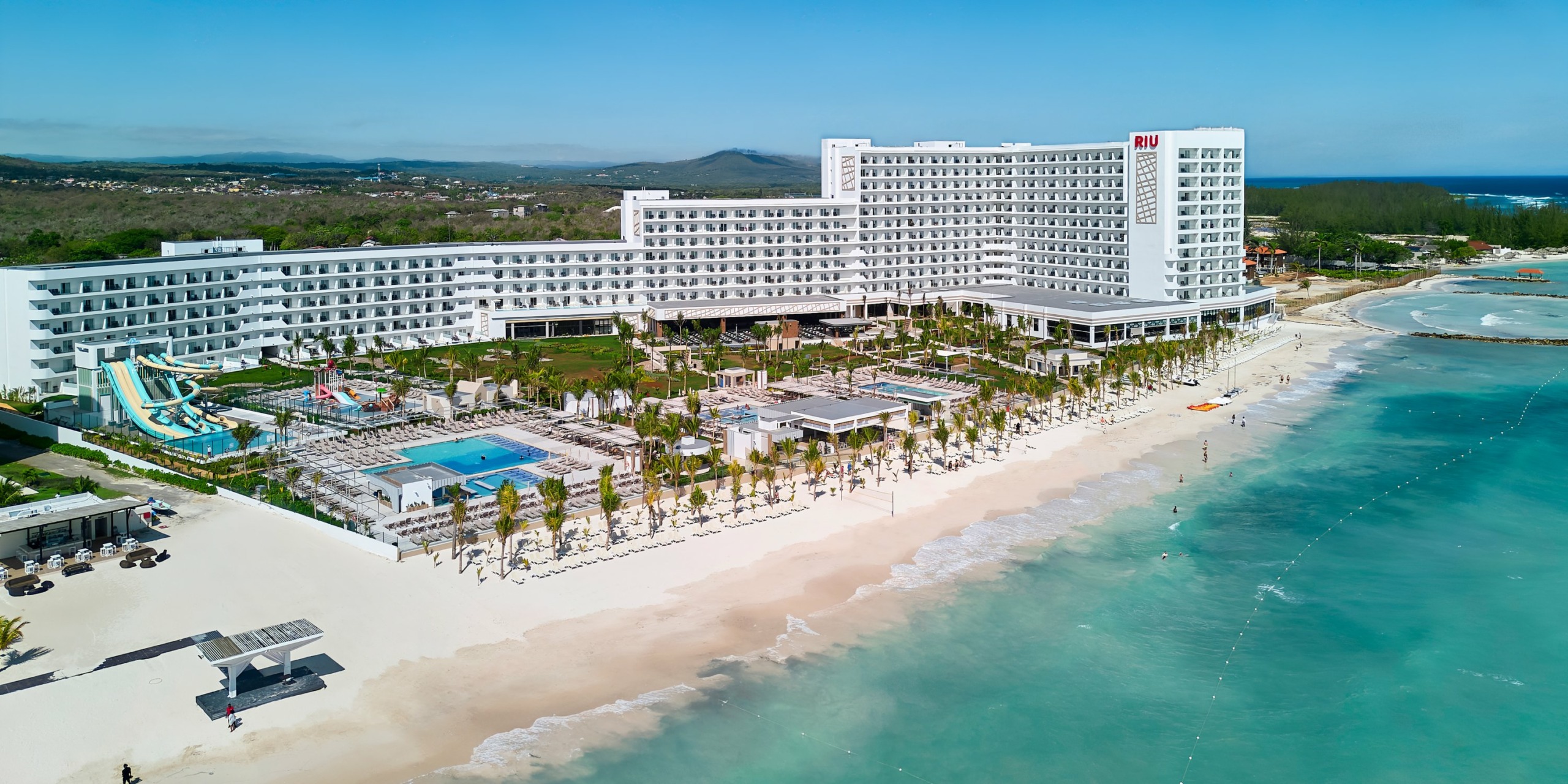 Aerial view of a large beachfront resort, RIU Palace Aquarelle, with multiple white buildings, swimming pools, water slides, sun loungers, and surrounding greenery. The sea is calm and clear, with some guests visible.