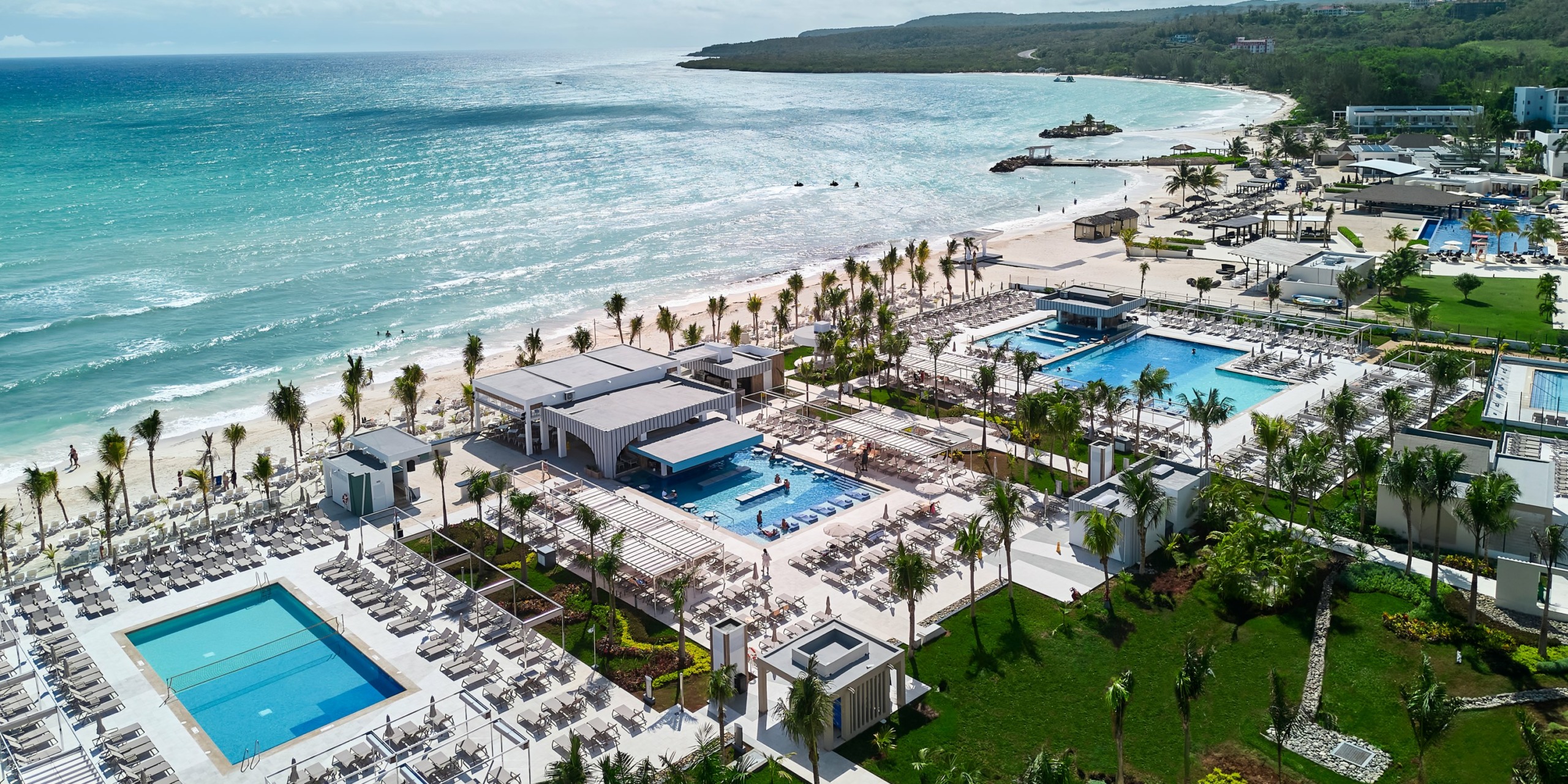 Aerial view of RIU Palace Aquarelle with multiple swimming pools, lounge chairs, and manicured gardens, set against a backdrop of the ocean and a distant shoreline.