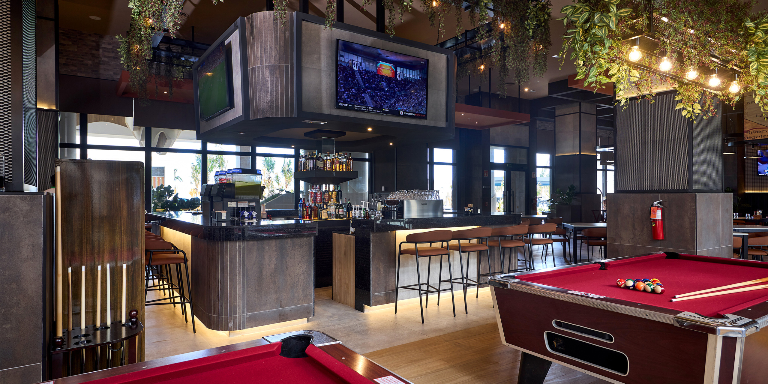 A modern bar area with a central counter, overhead TV, high stools, and a RIU Palace Aquarelle in the foreground. The space is well-lit with natural light and hanging plants decorating the interior.
