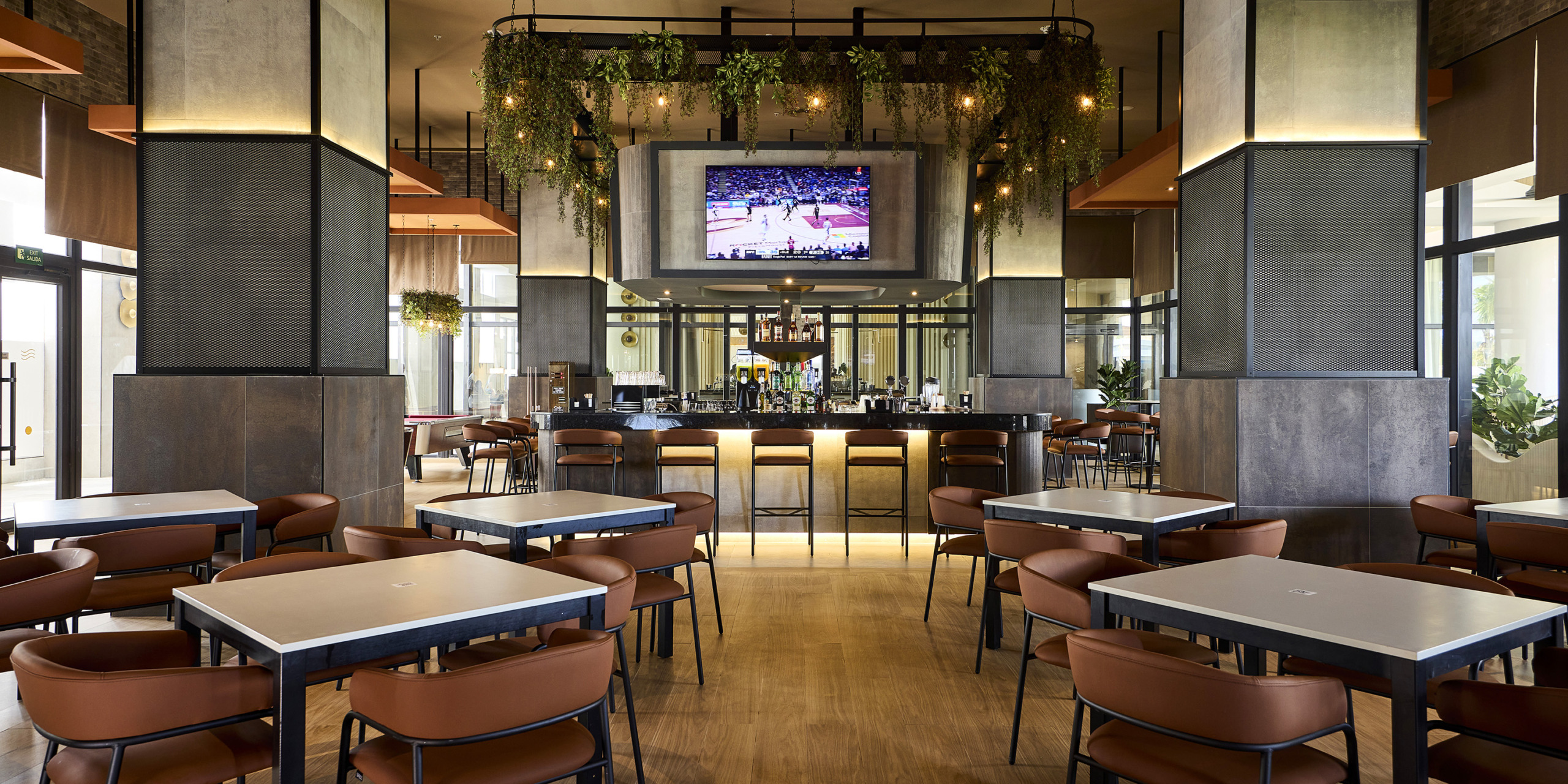 A modern bar with square tables and brown chairs, plants hanging from the ceiling, and a large screen in the background displaying RIU Palace Aquarelle.