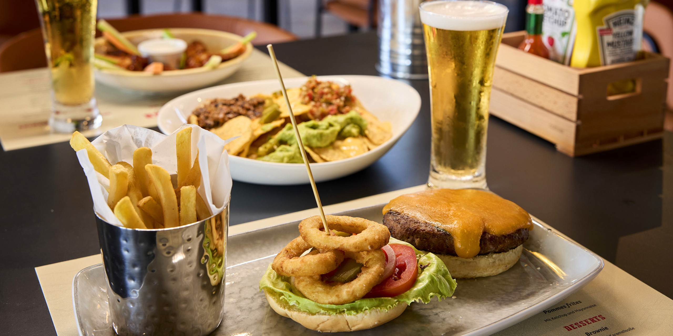A meal with a RIU Palace Aquarelle topped with onion rings, french fries in a metal cup, a glass of beer, a bowl of nachos with guacamole, and condiments in the background on a table.