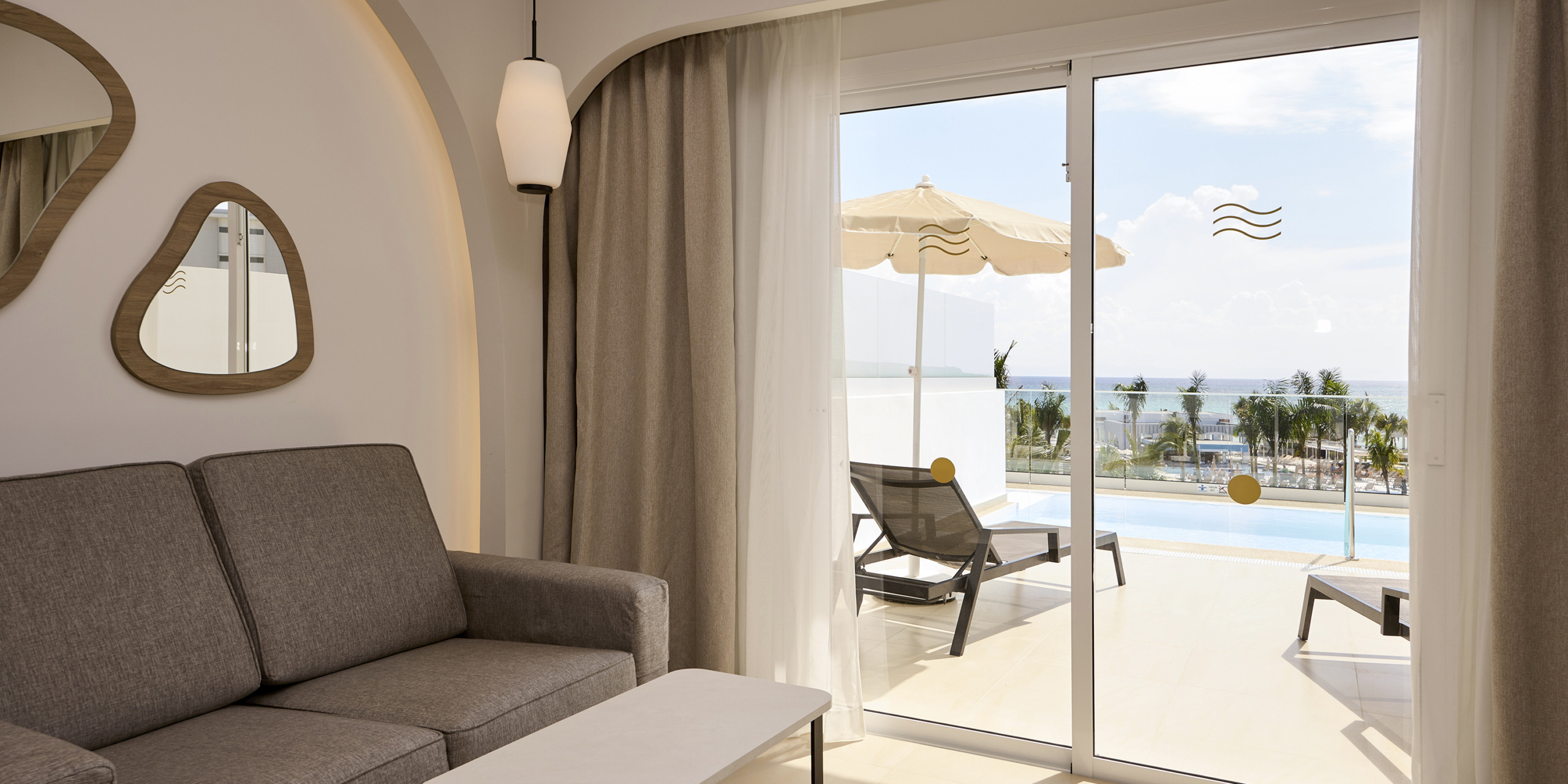 A modern hotel room with a gray sofa, white coffee table, and decorative mirrors. Sliding glass doors open to a balcony featuring lounge chairs, an umbrella, and a view of RIU Palace Aquarelle.