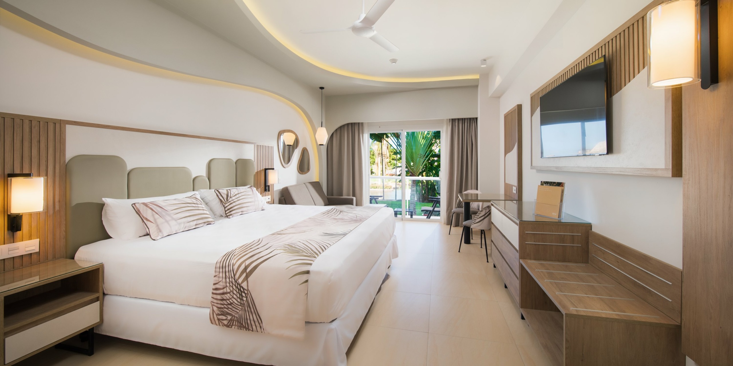 A modern hotel room with a large bed, wall-mounted TV, desk, and seating area at the RIU Palace Aquarelle. The space has light wood accents and a sliding door leading to an outdoor area with greenery.