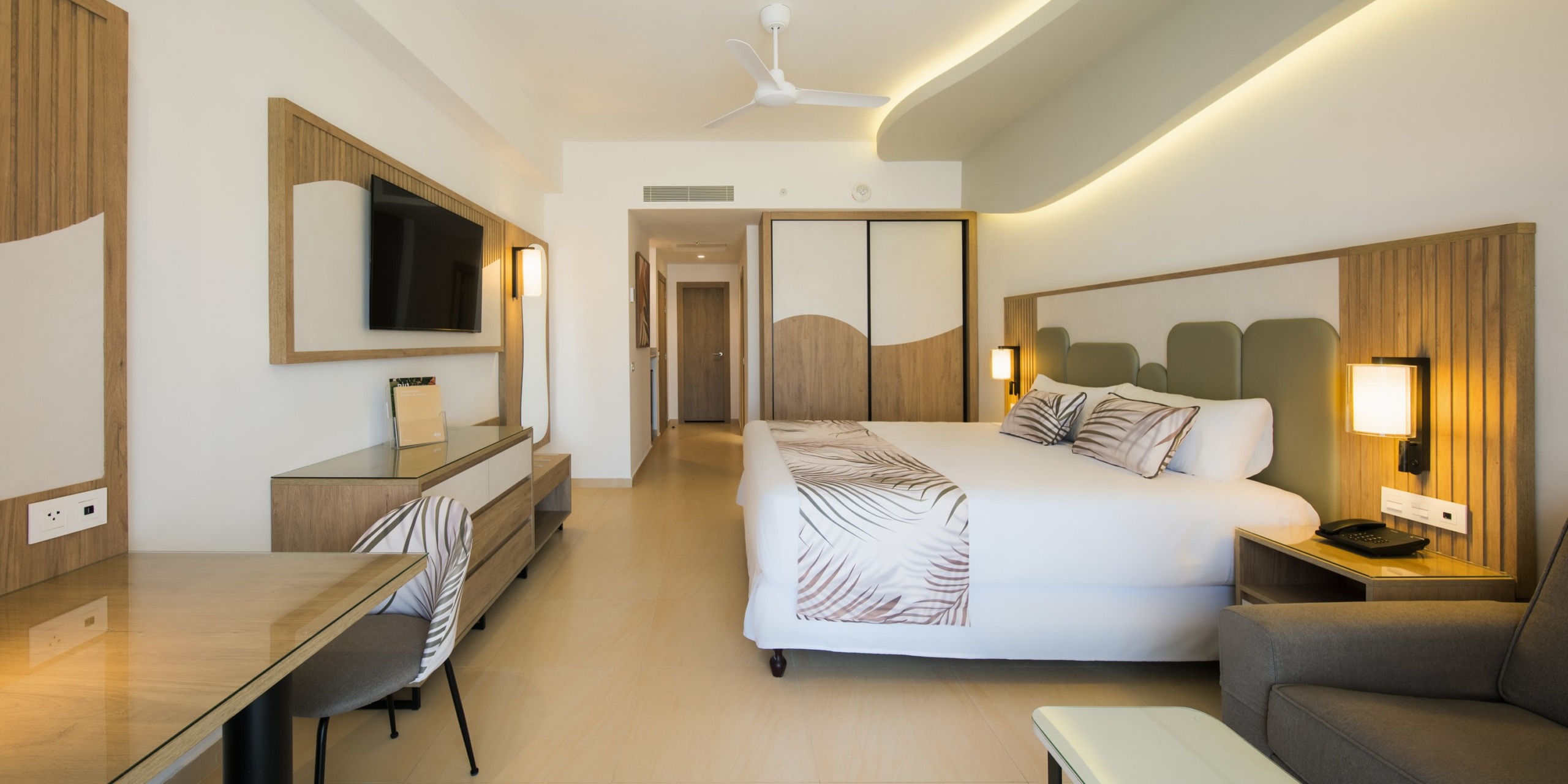 A modern hotel room with a large bed, desk, chair, flat-screen TV, and contemporary decor. The RIU Palace Aquarelle features wooden accents, soft lighting, and a ceiling fan.
