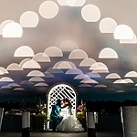 A bride and groom sit closely under a white archway decorated with flowers at night, with an artistic overlay of illuminated semicircles in the sky and boats in the background.