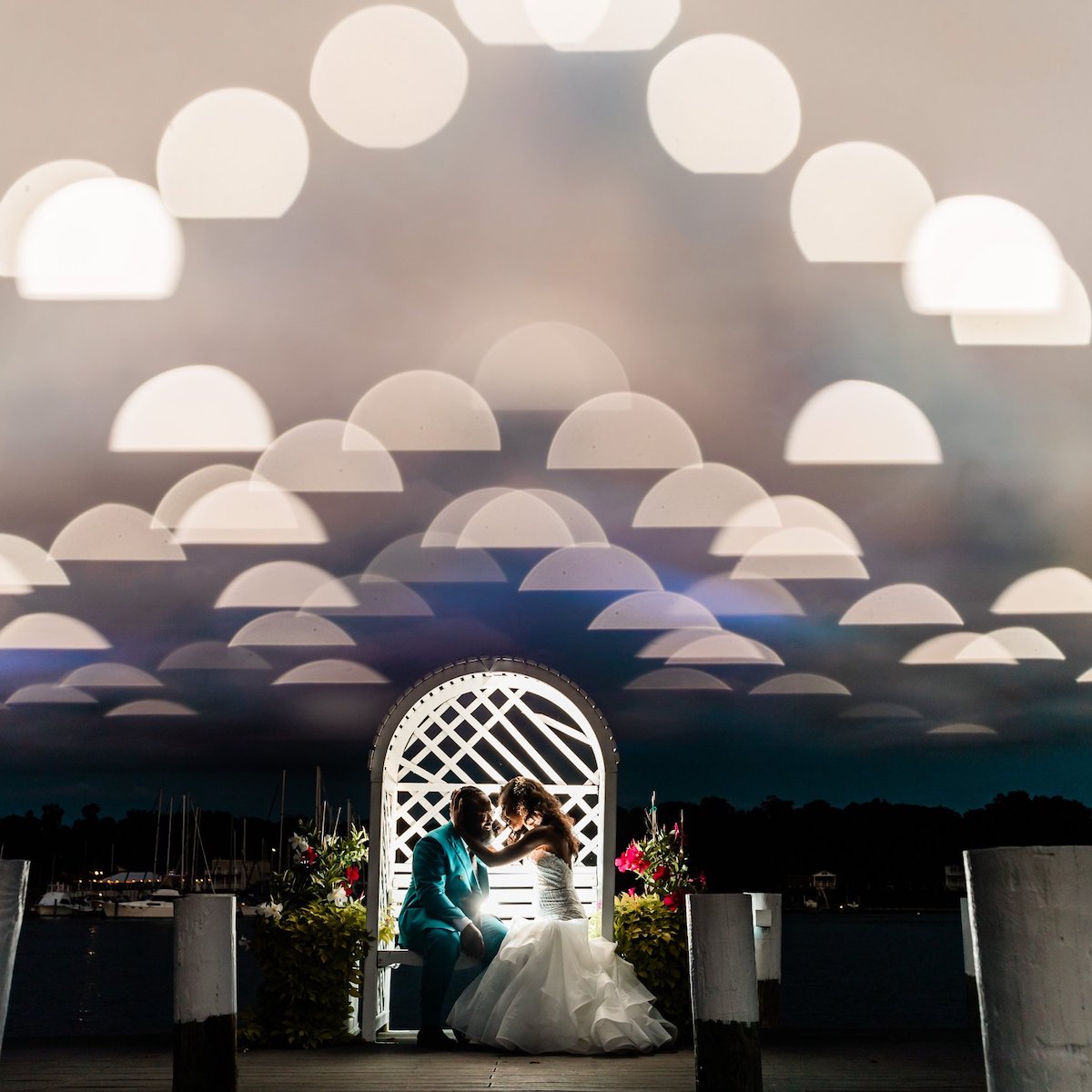 A bride and groom sit closely under a white archway decorated with flowers at night, with an artistic overlay of illuminated semicircles in the sky and boats in the background.