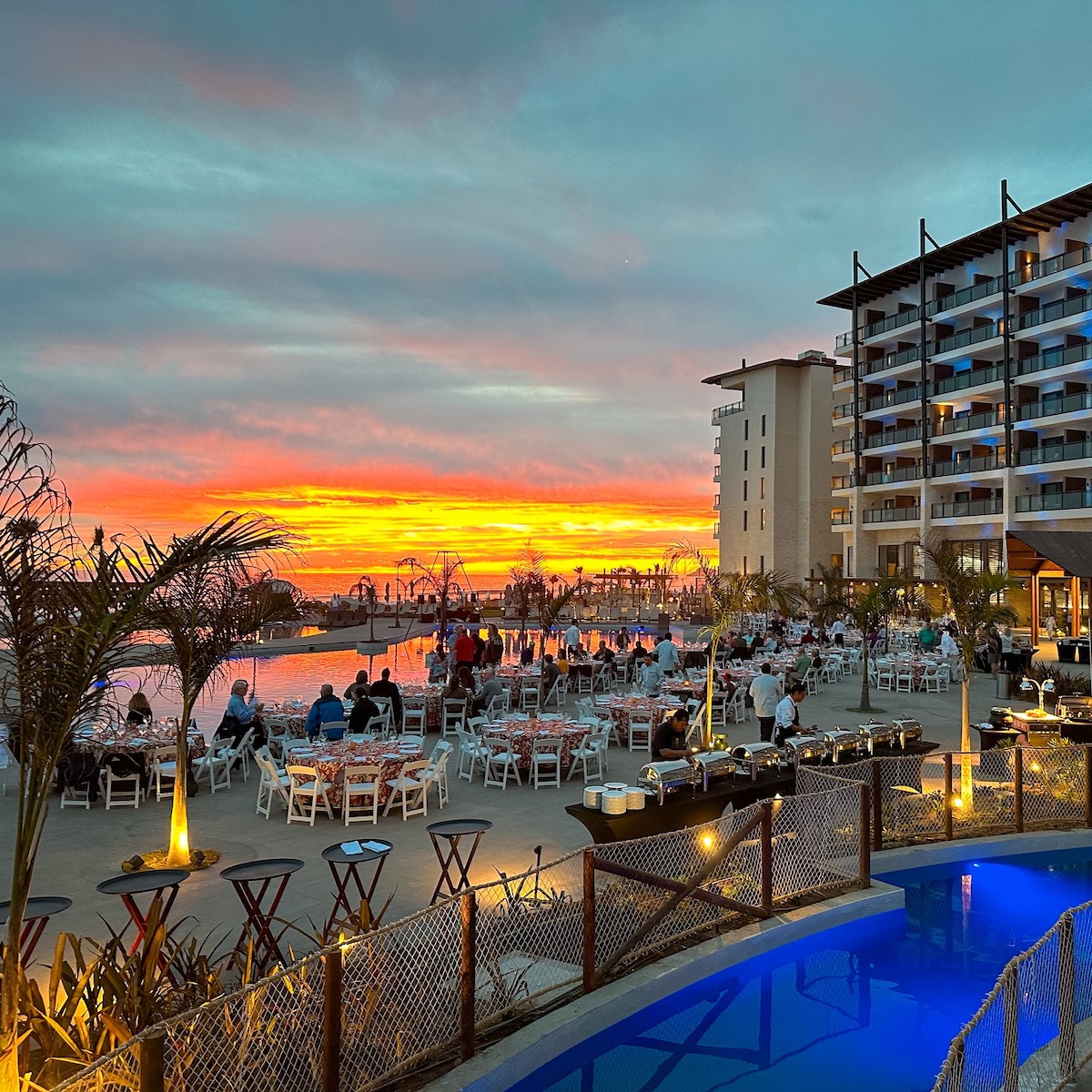 View of the sunset over Dreams Estrella del Mar Mazatlan resort, featuring tables and chairs arranged near a pool. Guests are seated, taking in the beautiful surroundings, with a multi-story building on the right beneath a vibrant sky.