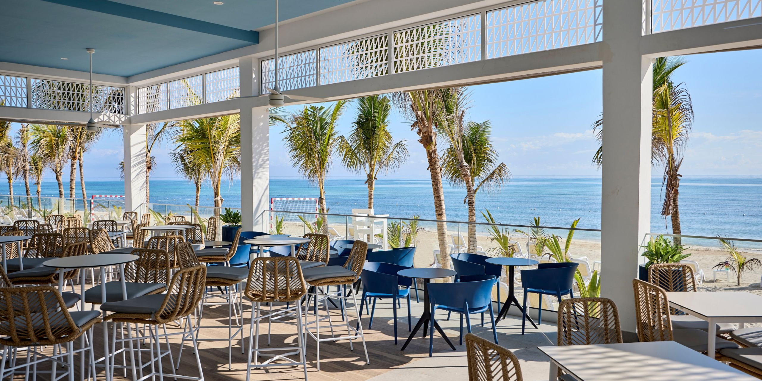 Hotel RIU Palace Aquarelle features an open-air beachside café with tables and chairs, offering a stunning view of the ocean and palm trees beneath a clear sky.