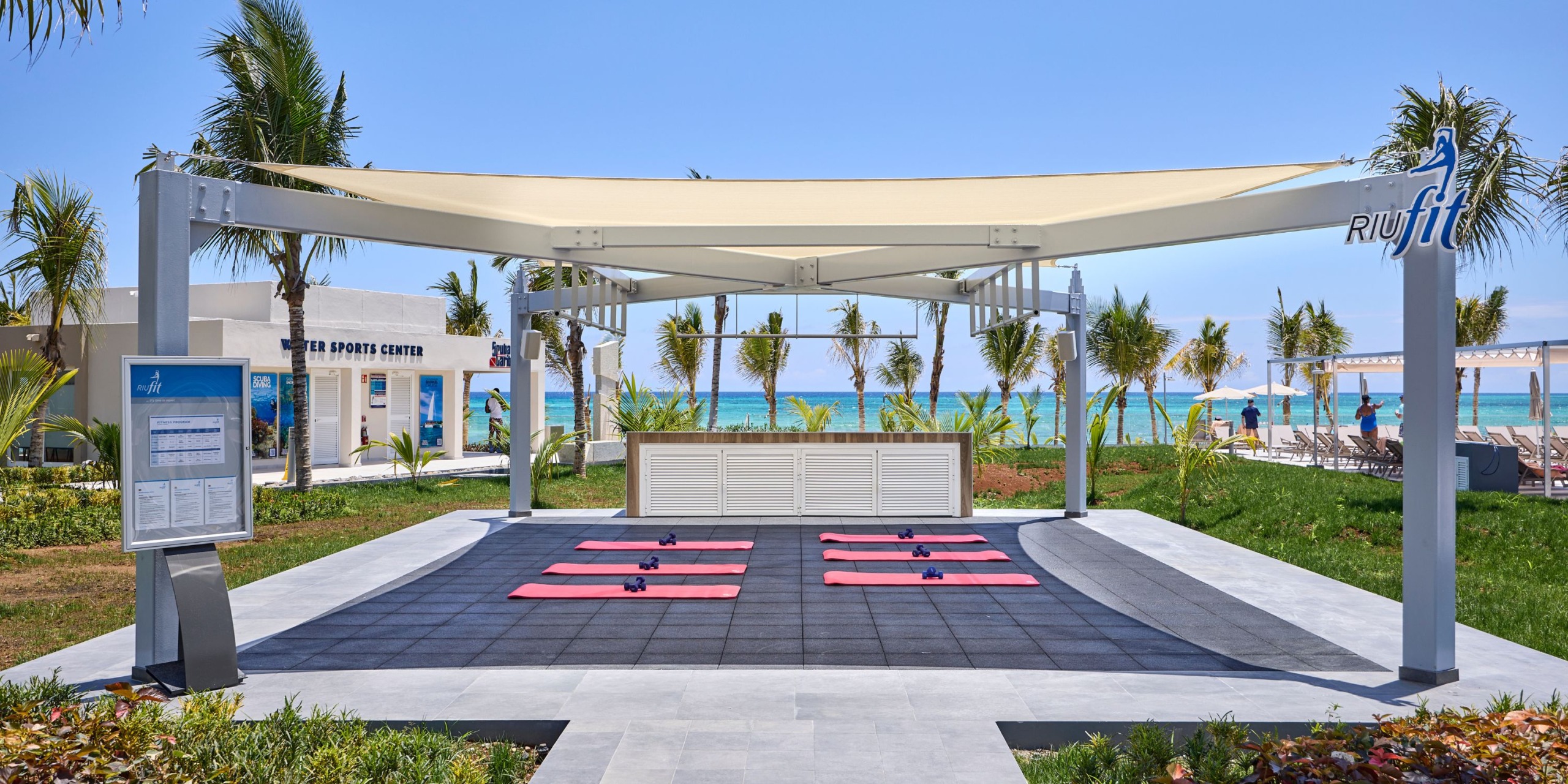 Experience the outdoor fitness area at Hotel RIU Palace Aquarelle, where yoga mats and dumbbells are available under a sunshade, with a breathtaking view of palm trees and the ocean.