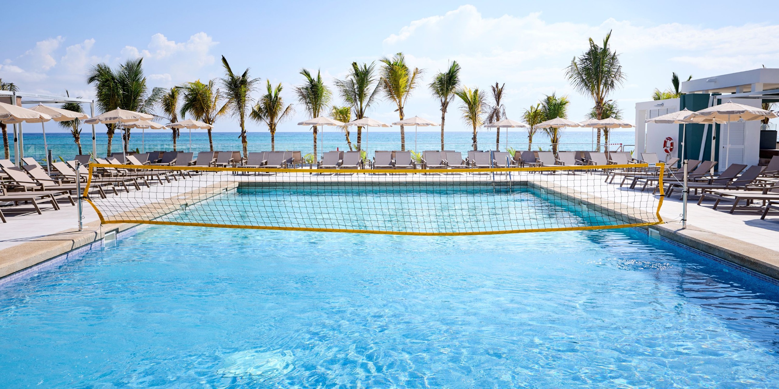 The Hotel RIU Palace Aquarelle offers a swimming pool featuring a volleyball net, surrounded by lounge chairs and palm trees, with an ocean view.