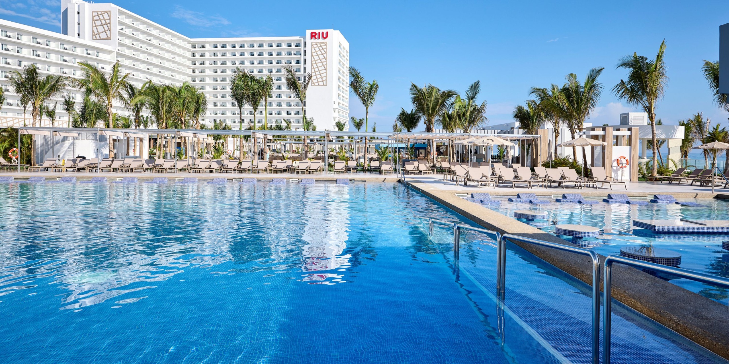 The Hotel RIU Palace Aquarelle features a pool area complete with loungers, umbrellas, and palm trees encircling the space. In the background, you can see the multi-story hotel building standing under a clear blue sky.