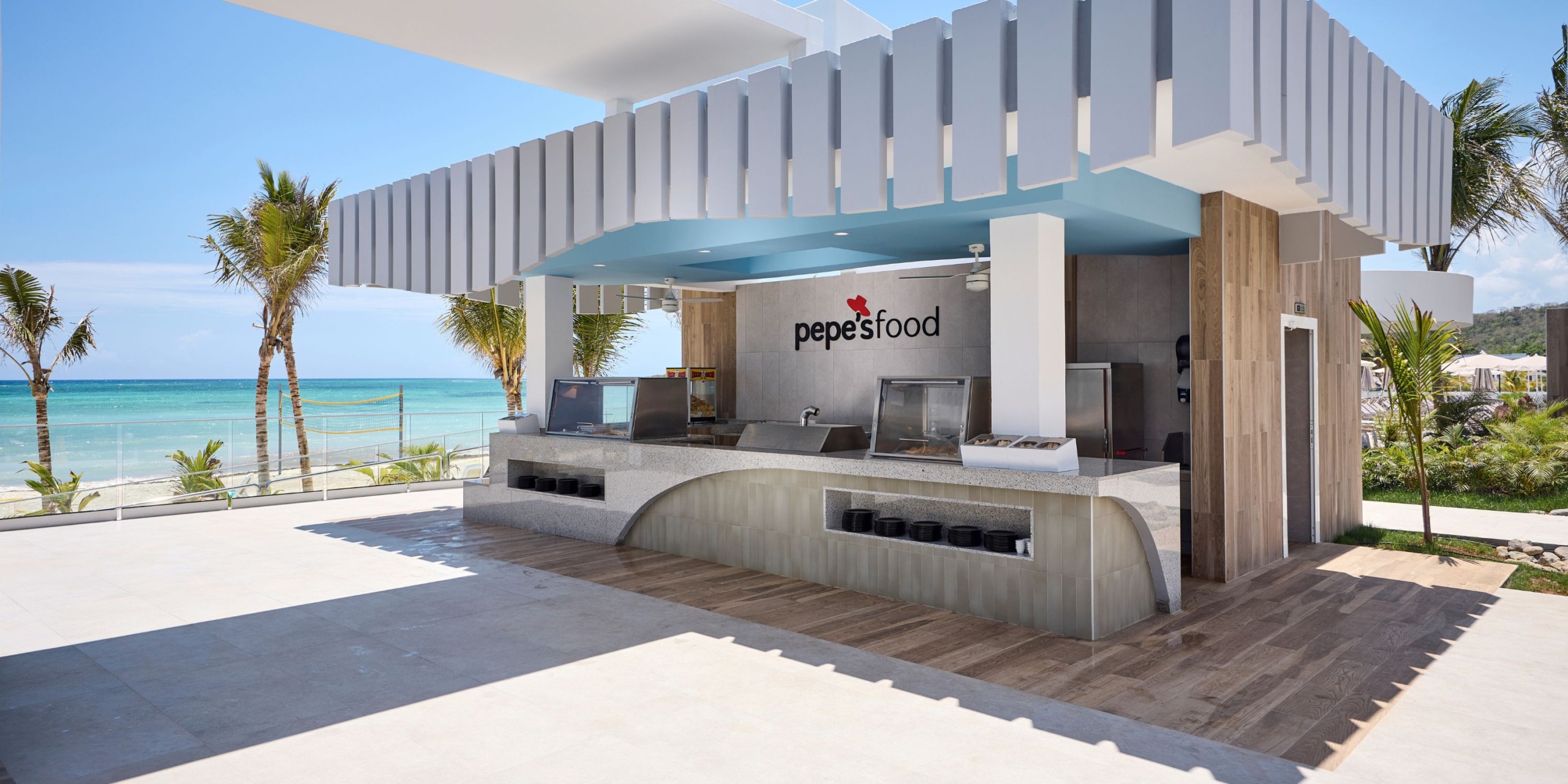 Outdoor beachside food stand at Hotel RIU Palace Aquarelle, labeled "pepe's food," featuring cooking equipment and surrounded by palm trees, with a view of the ocean.