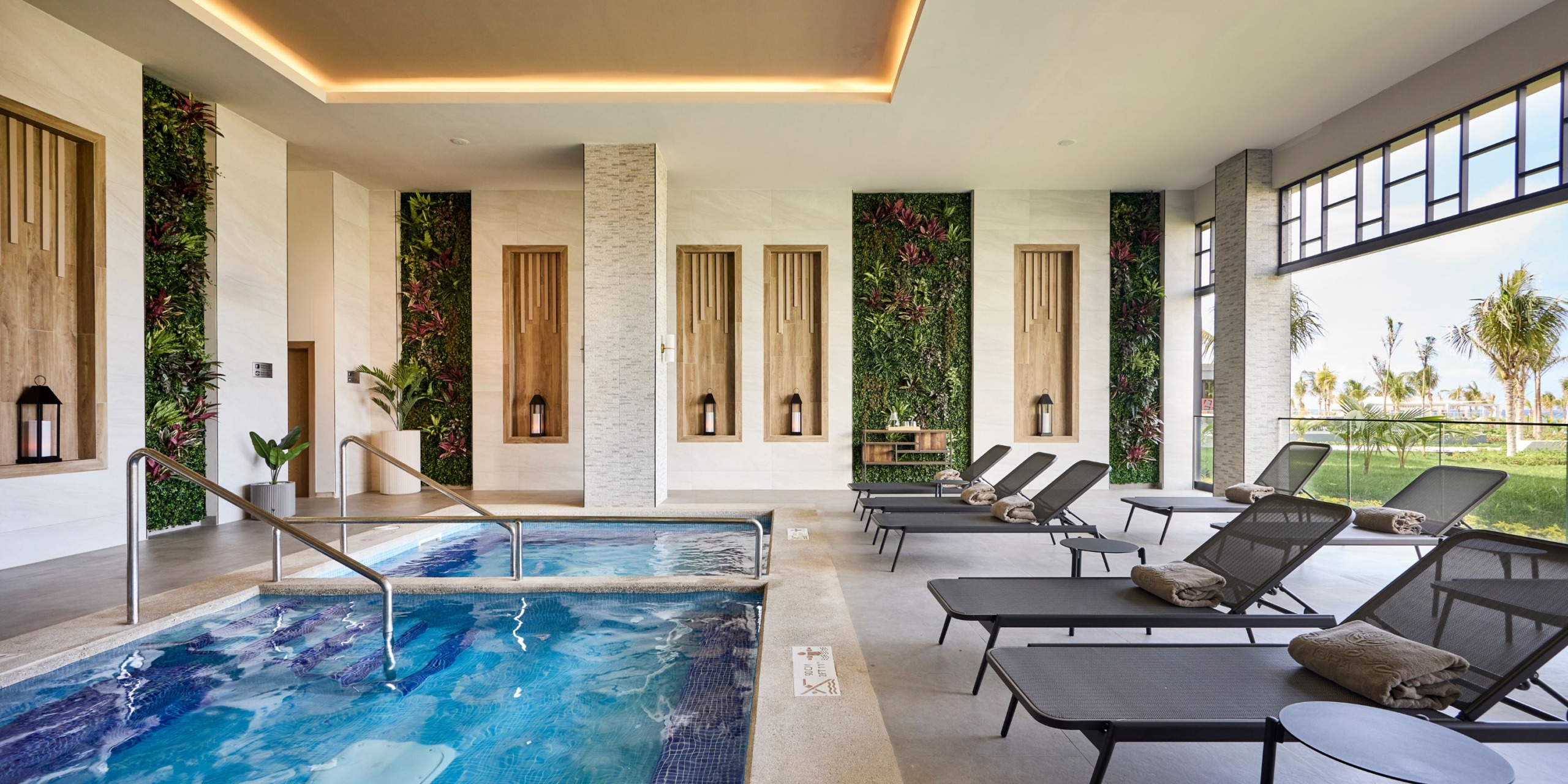 The indoor spa area at Hotel RIU Palace Aquarelle boasts decorative walls and includes a pool, loungers, and folded towels, with large windows offering views of the palm trees outside.