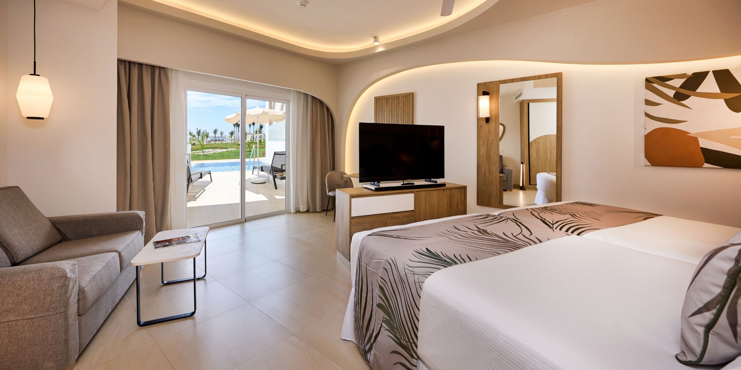 Hotel RIU Palace Aquarelle room featuring two beds, a sofa, and a flat-screen TV. The space is enhanced by large windows that open onto a patio with views of the pool and outdoor seating area. The room is beautifully finished with neutral tones and decor.